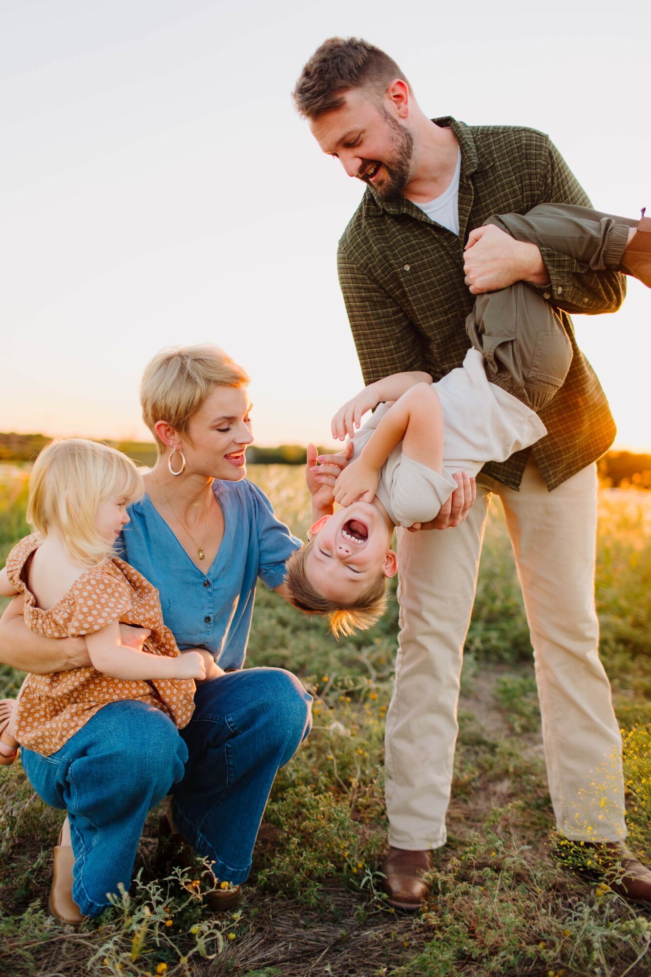 Family outdoors with a man lifting a child, woman and another child sitting on the ground smiling.