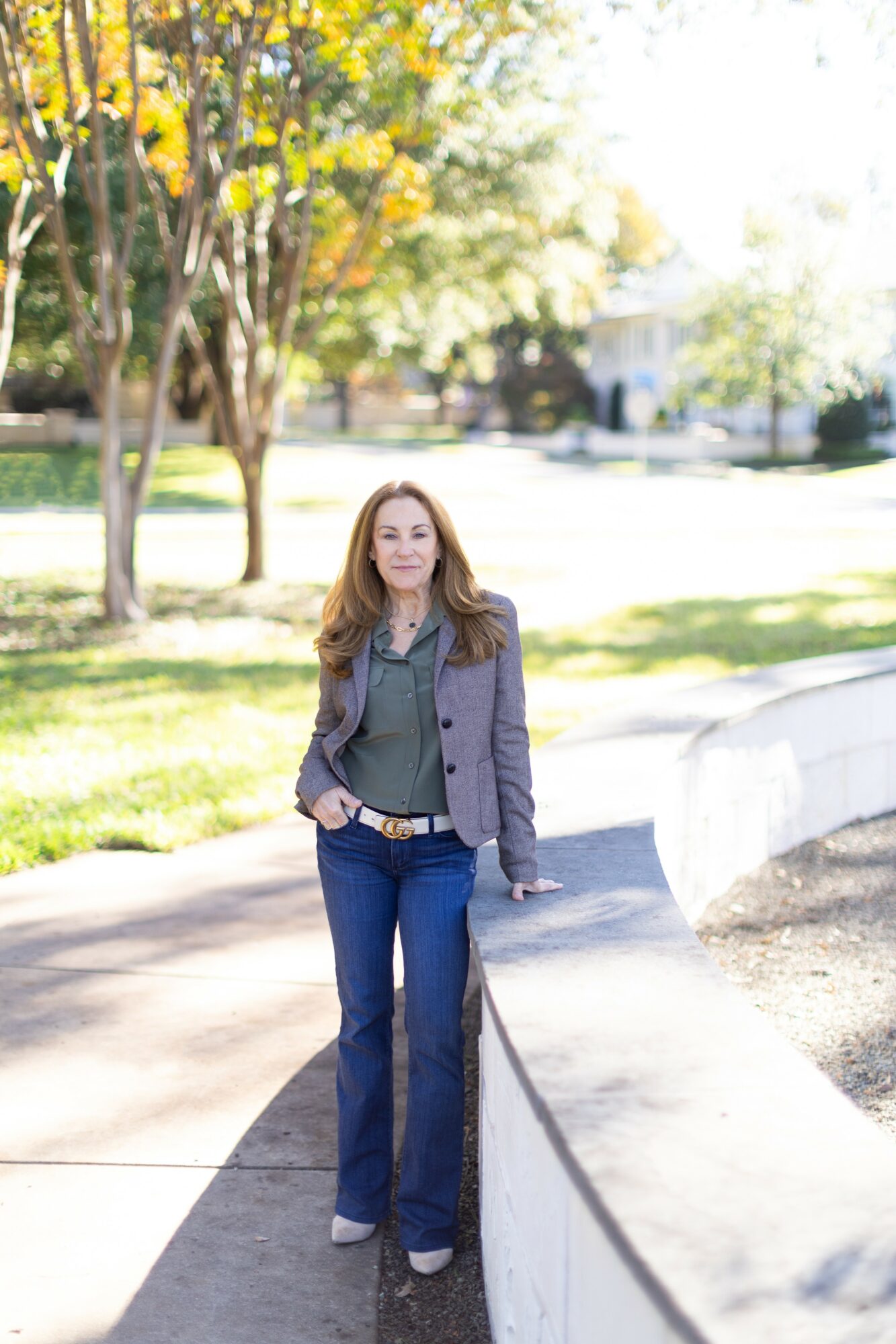 Woman standing outdoors on a sidewalk with trees and greenery in the background.