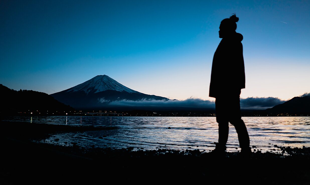 Person standing near water with mountain and snow-capped peak in background during dusk or dawn.