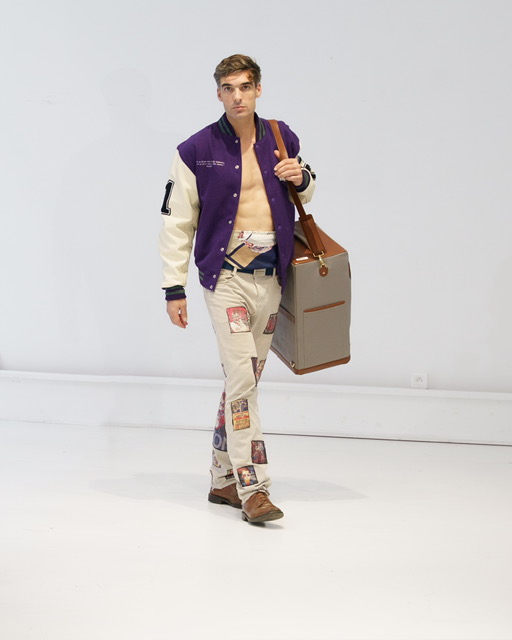 Young man walking with a large bag, wearing a varsity jacket and patched pants, against a plain white background.