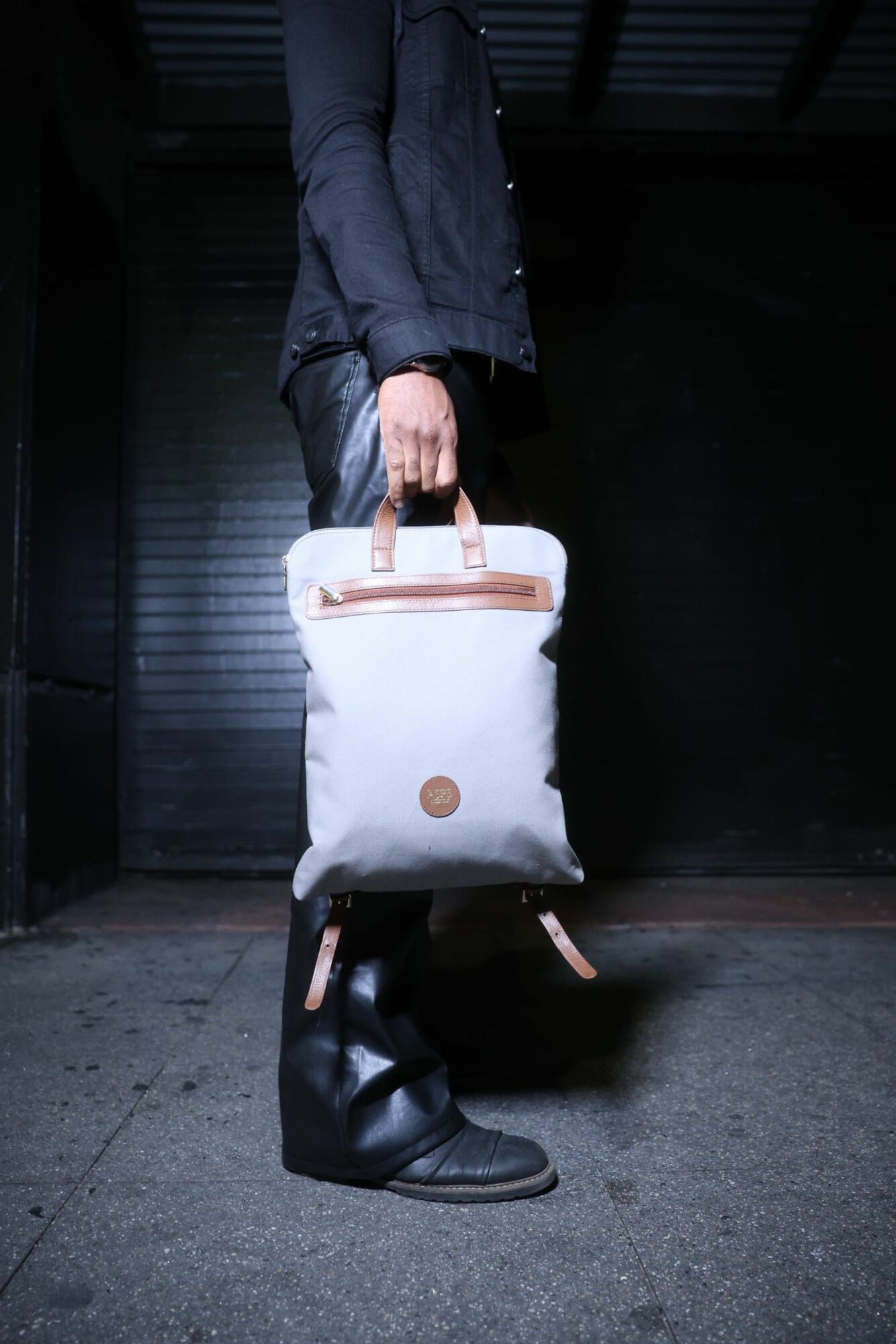 Person standing in dark environment holding a white bag with brown handles and a round logo, wearing black shoes and dark clothing.