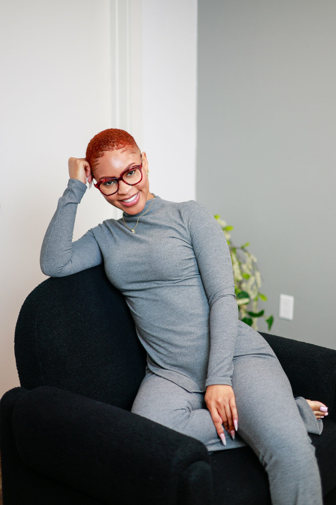 Woman with red hair and glasses sitting on a black couch, smiling, in a room with white and gray walls.