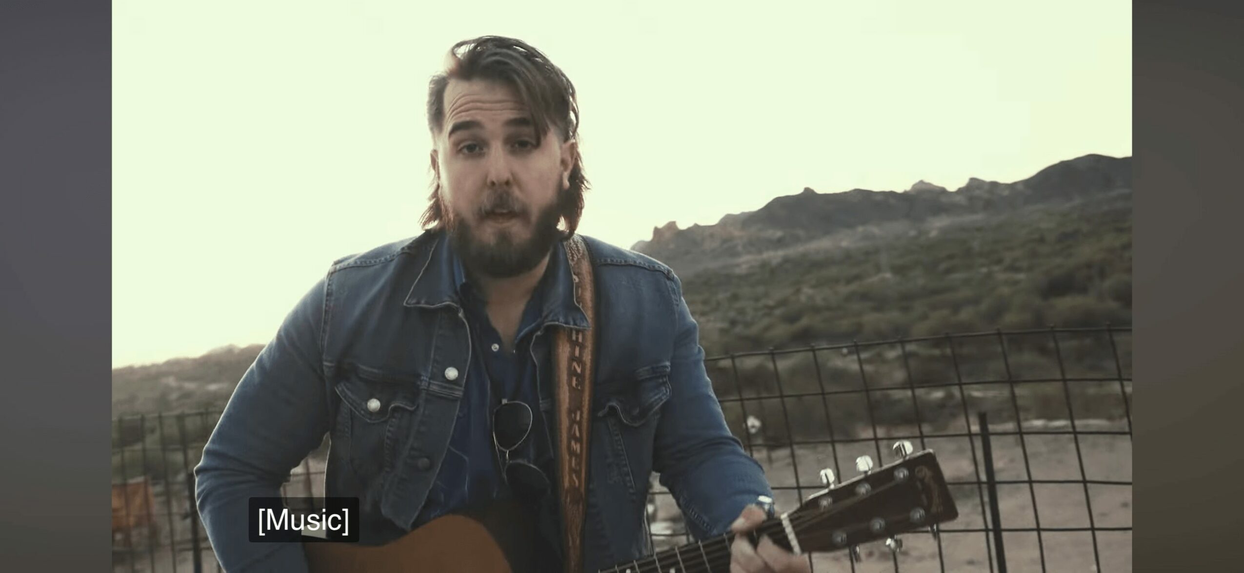 Man with beard and long hair playing guitar outdoors near a fence and hillside.