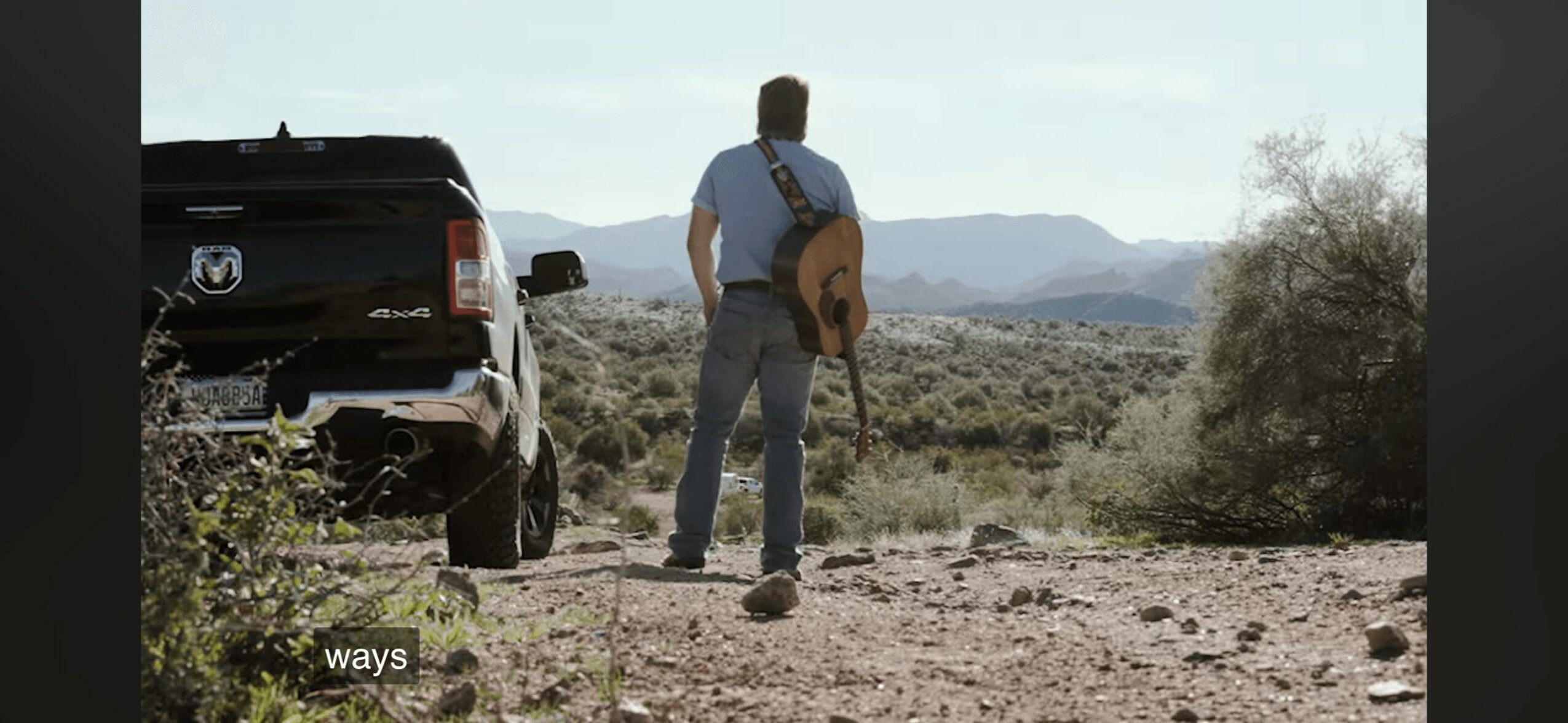 Person with a guitar case walking away from a vehicle in a desert landscape with mountains in the background.