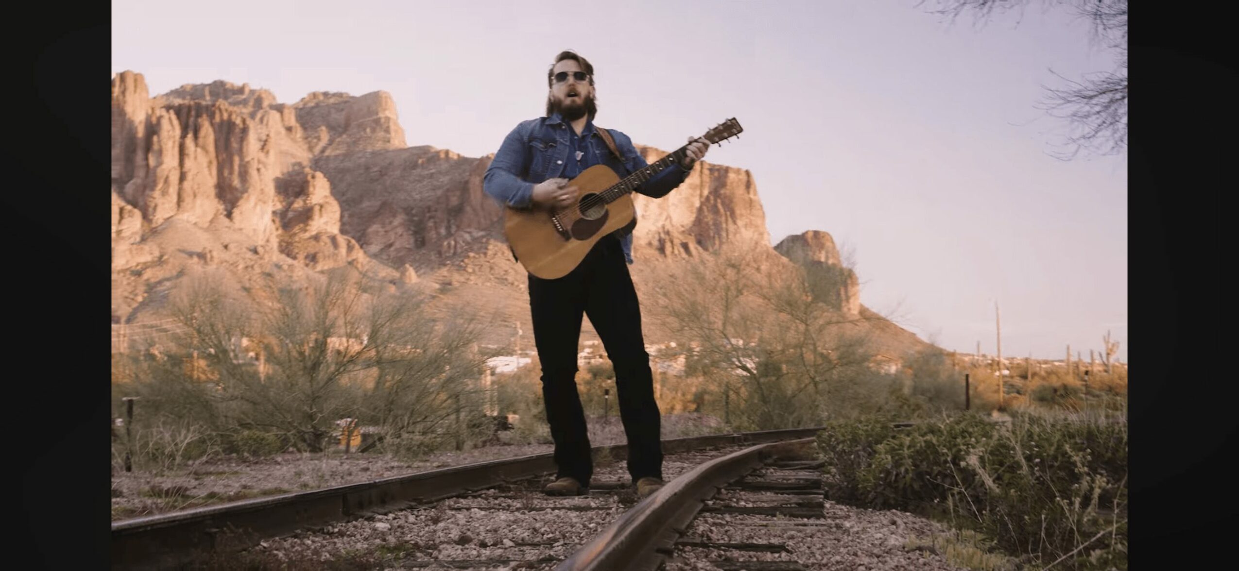 Man playing guitar on railway tracks with desert landscape and cliffs in background.