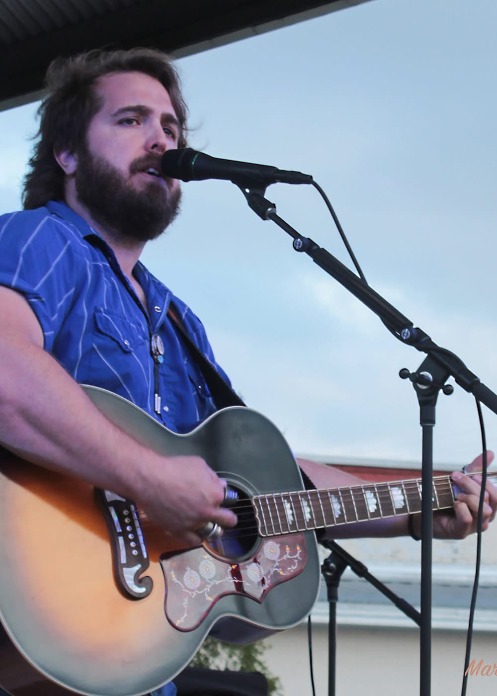 Man with beard playing guitar and singing into microphone outdoors.