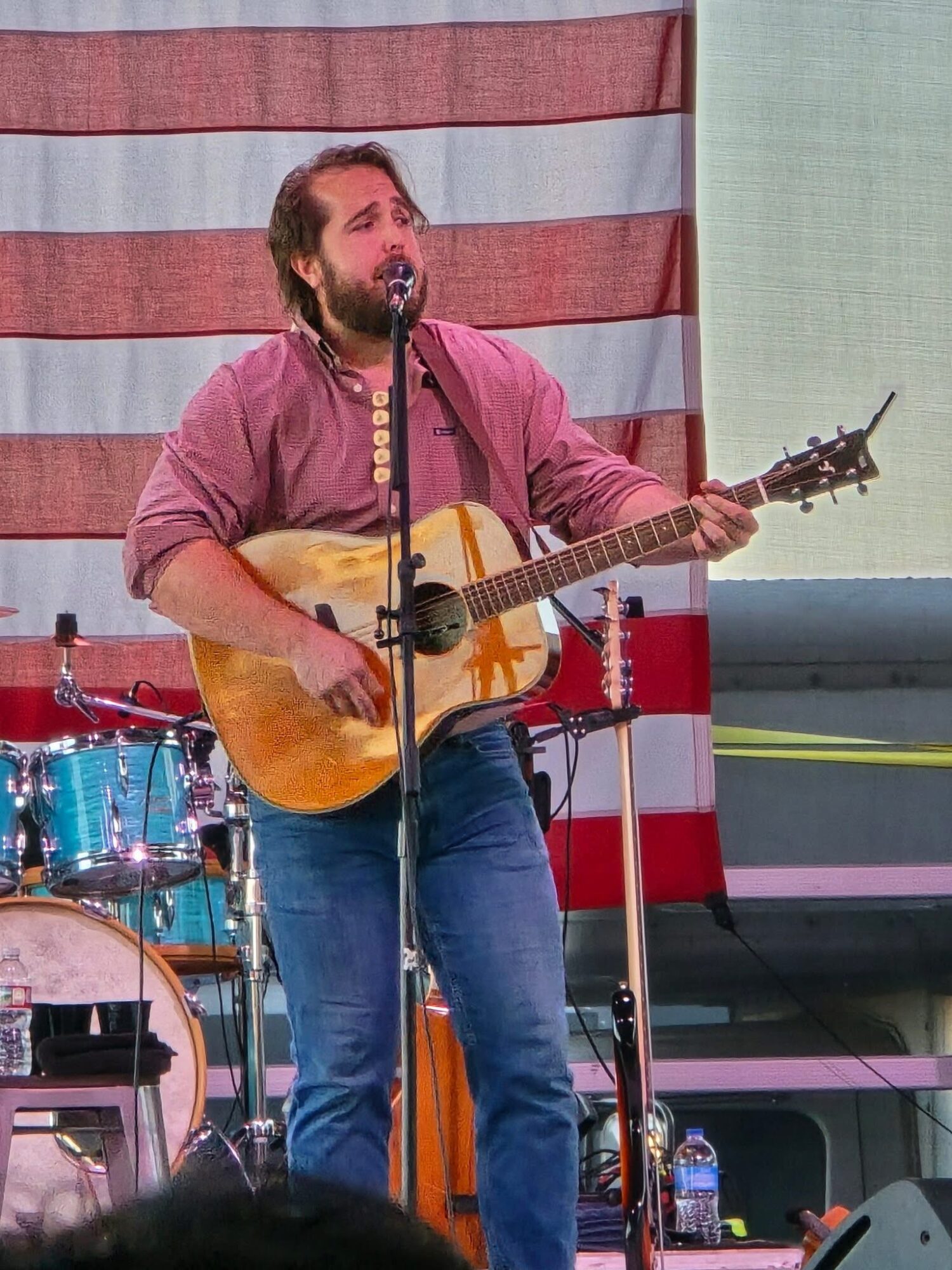 Man with beard and long hair playing an acoustic guitar on stage, with drums and American flag backdrop.