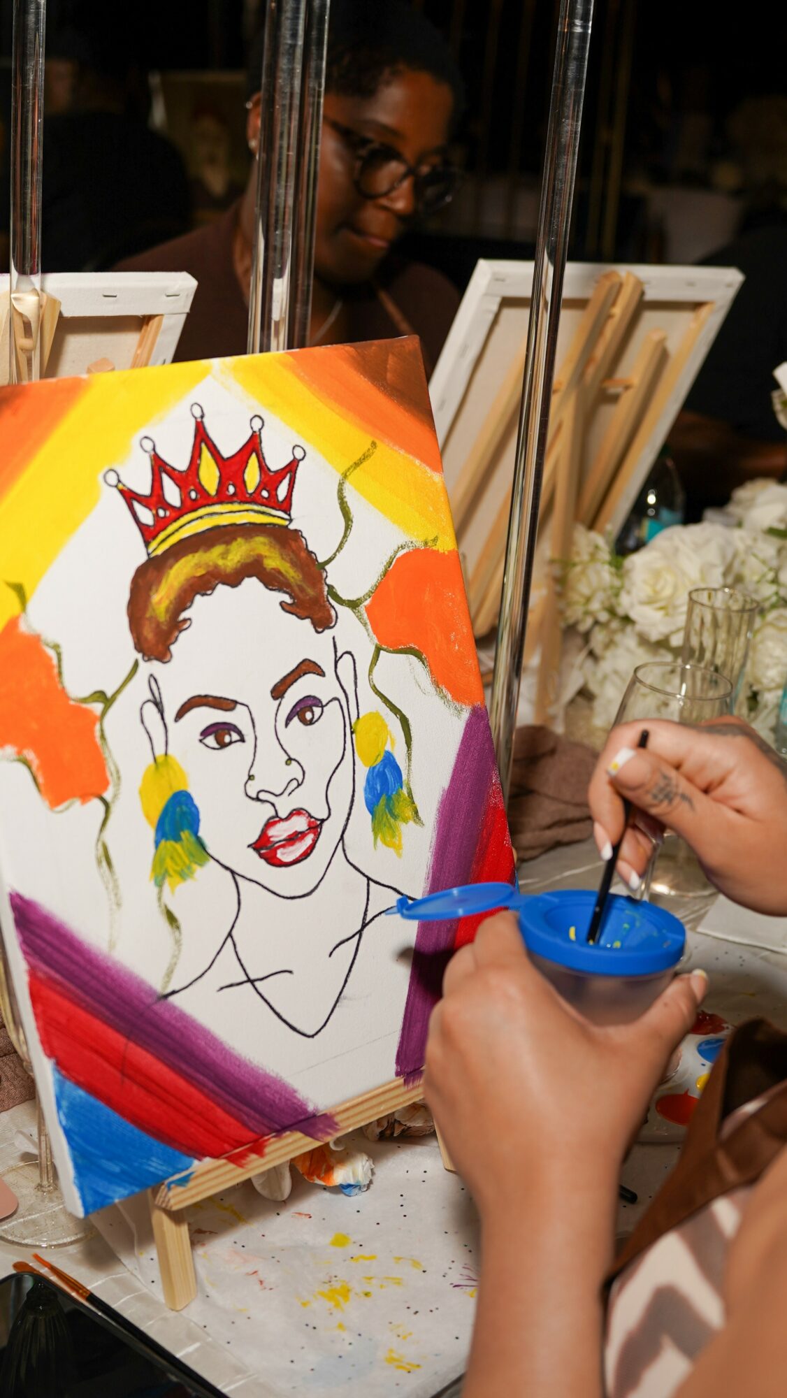 Person painting a colorful portrait of a woman with a crown on canvas, with art supplies nearby.