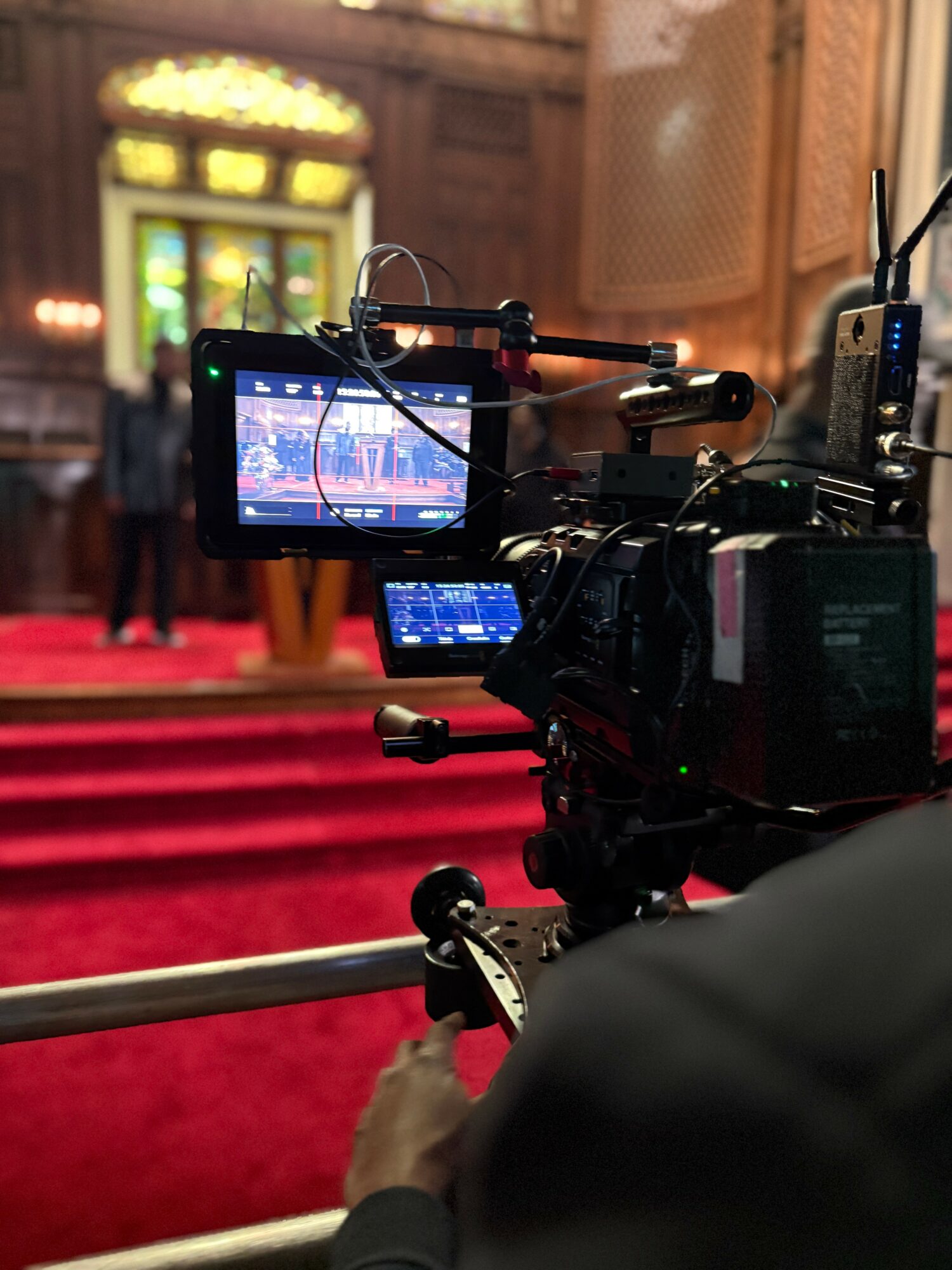 Camera on tripod filming a stage with red carpet and ornate background, focus on camera display screen.