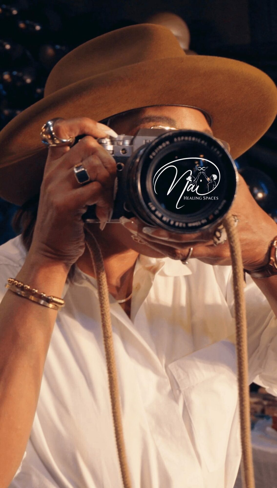 Person wearing a wide-brimmed hat taking a photo with a camera, wearing jewelry and a white shirt.