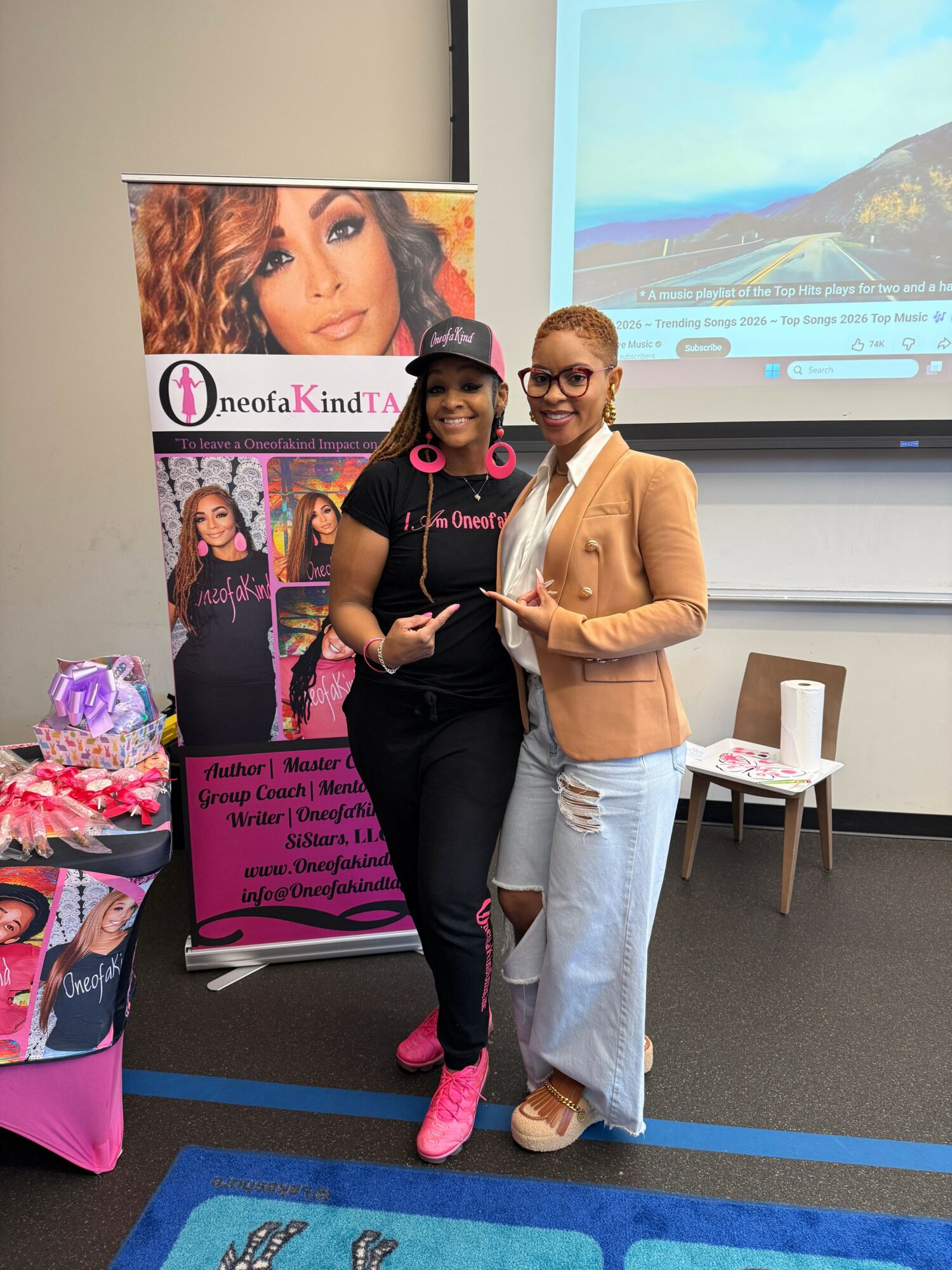 Two women standing together in a room with a presentation screen and a pink promotional banner behind them.