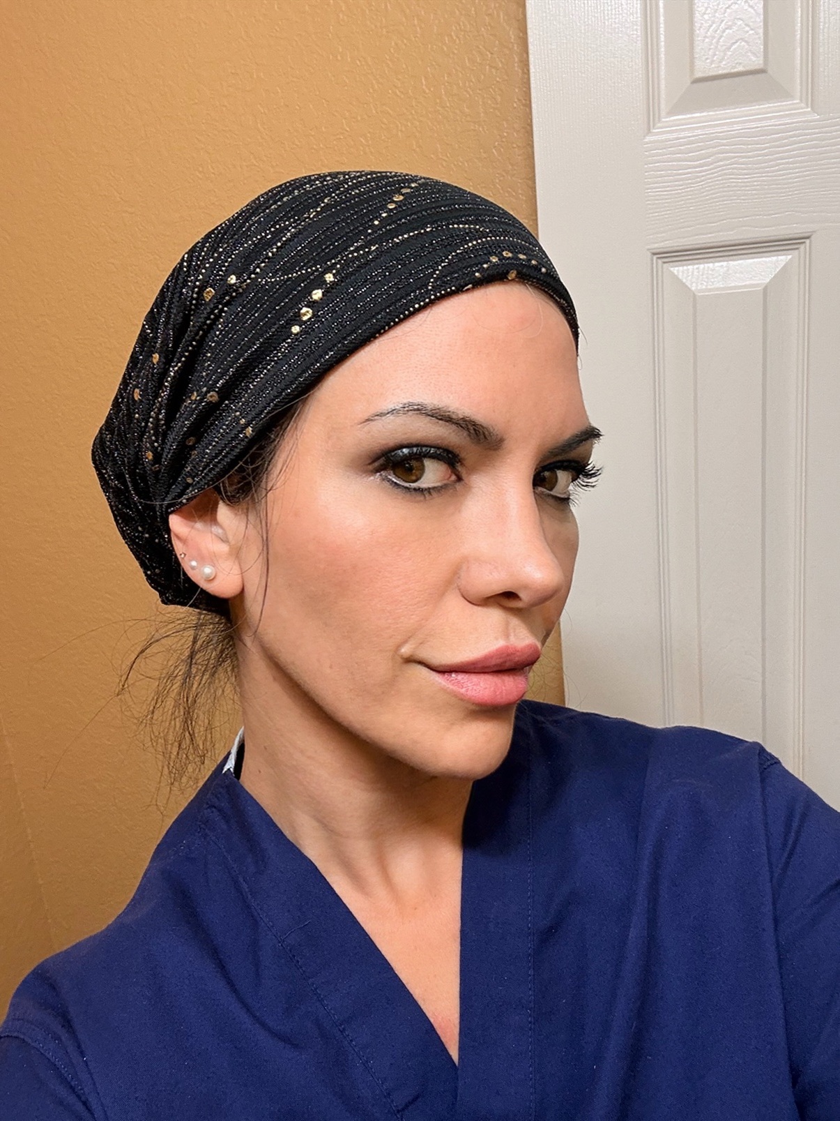 Woman with dark headscarf and blue top, looking at camera, in front of white door and beige wall.