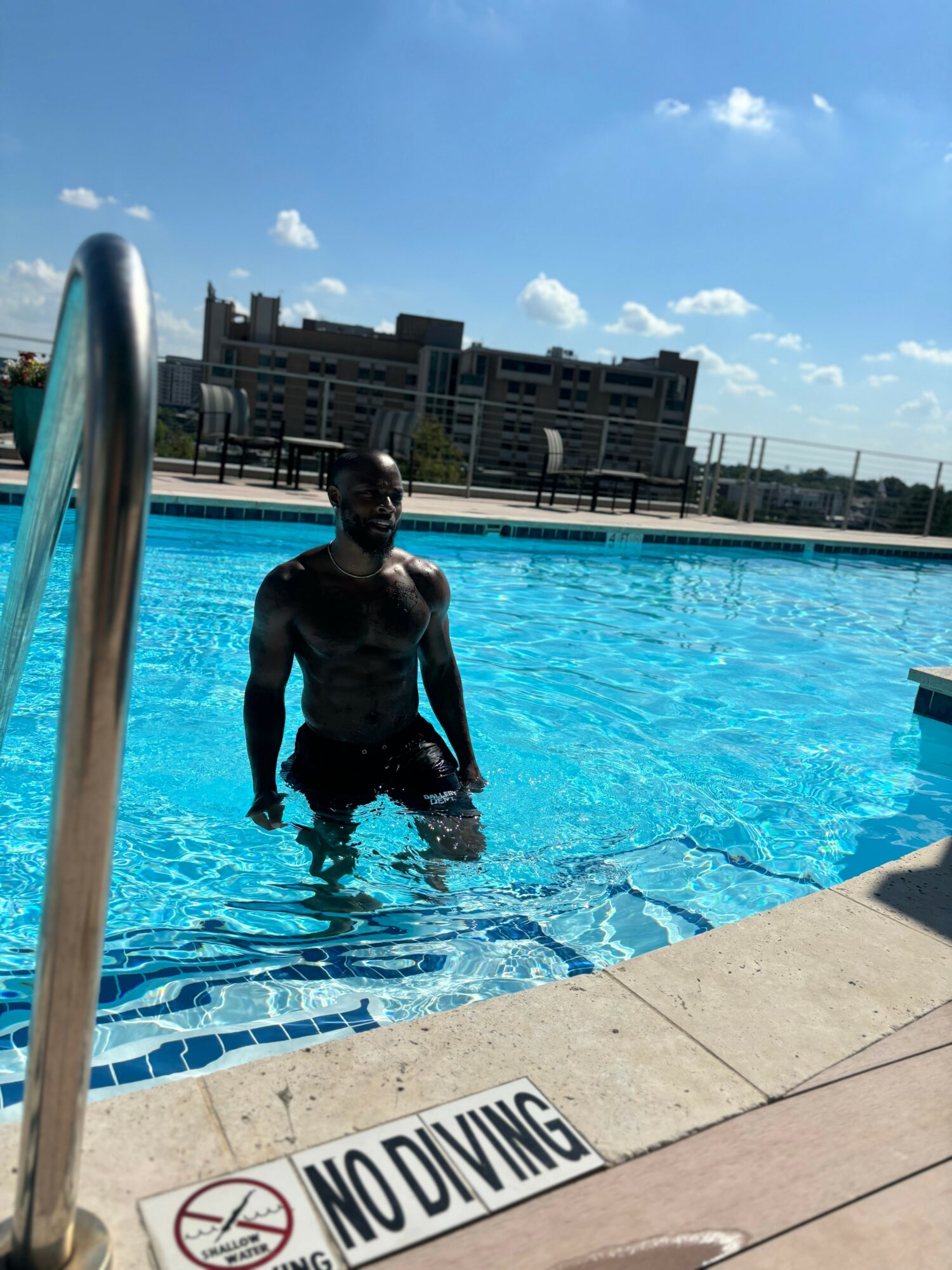 Person standing in a swimming pool near a ladder, with a building and blue sky in the background.