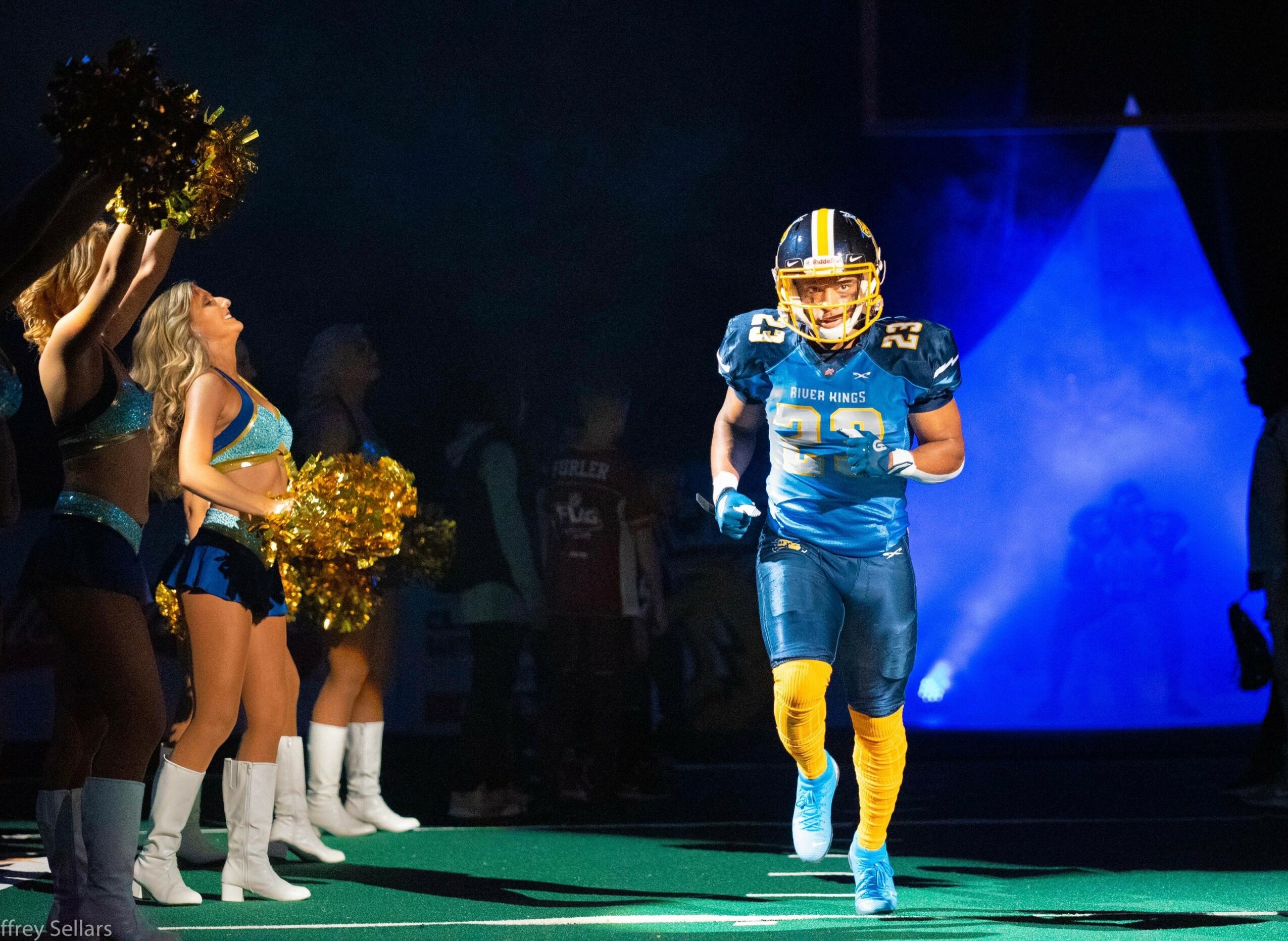 Football player in blue uniform running onto field with cheerleaders holding pom-poms nearby.