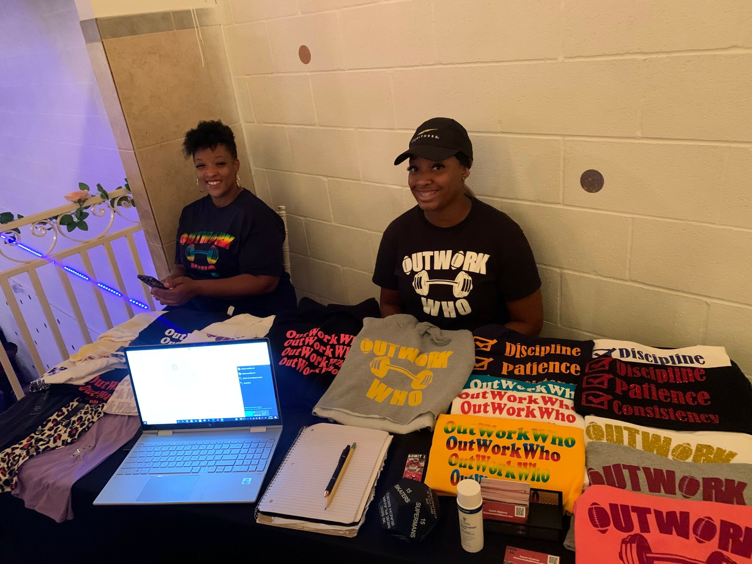 Two children sit at a table with laptops, books, and supplies in a room with beige walls.