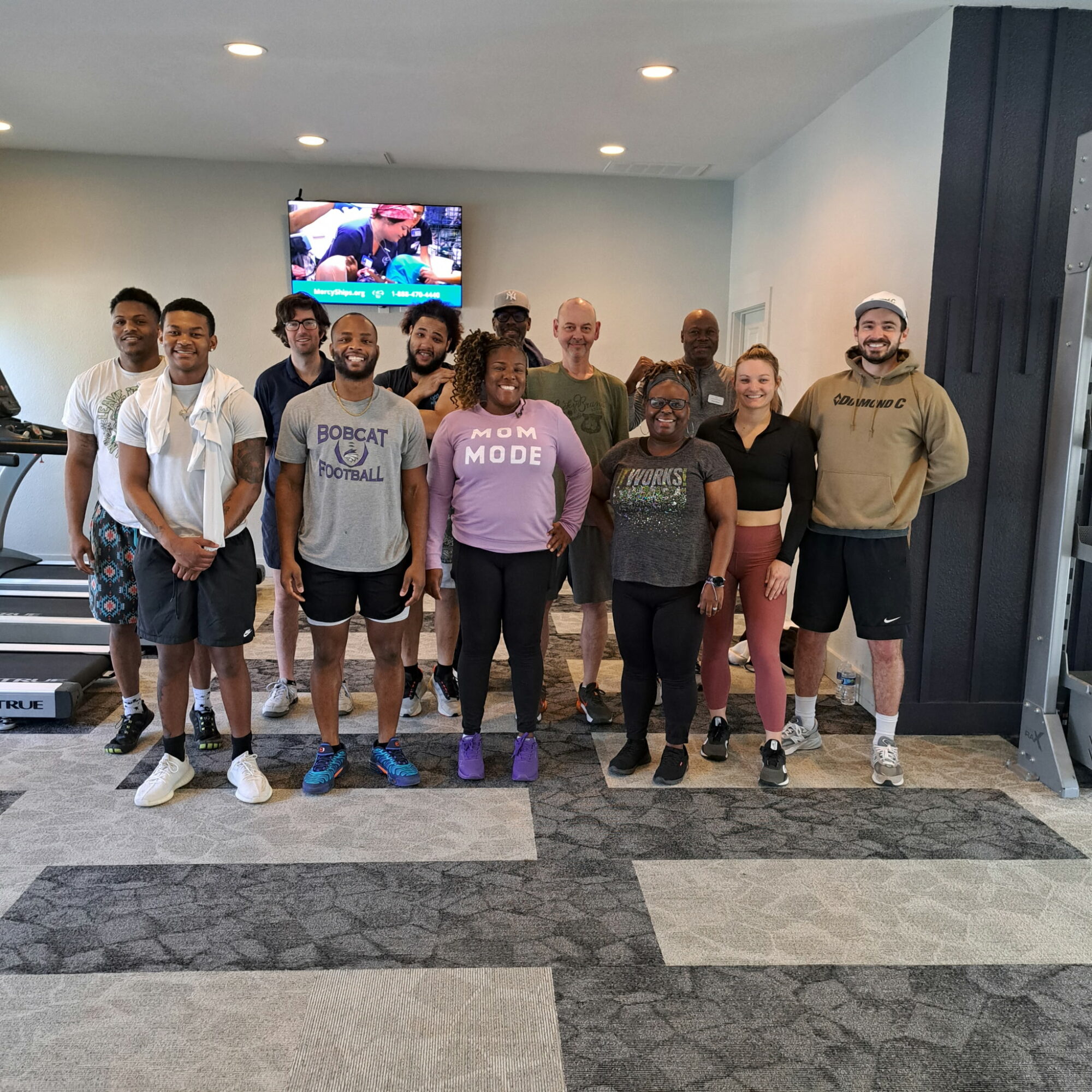 Group of 12 diverse people standing indoors, some smiling, in front of a wall with a TV screen, casual attire.