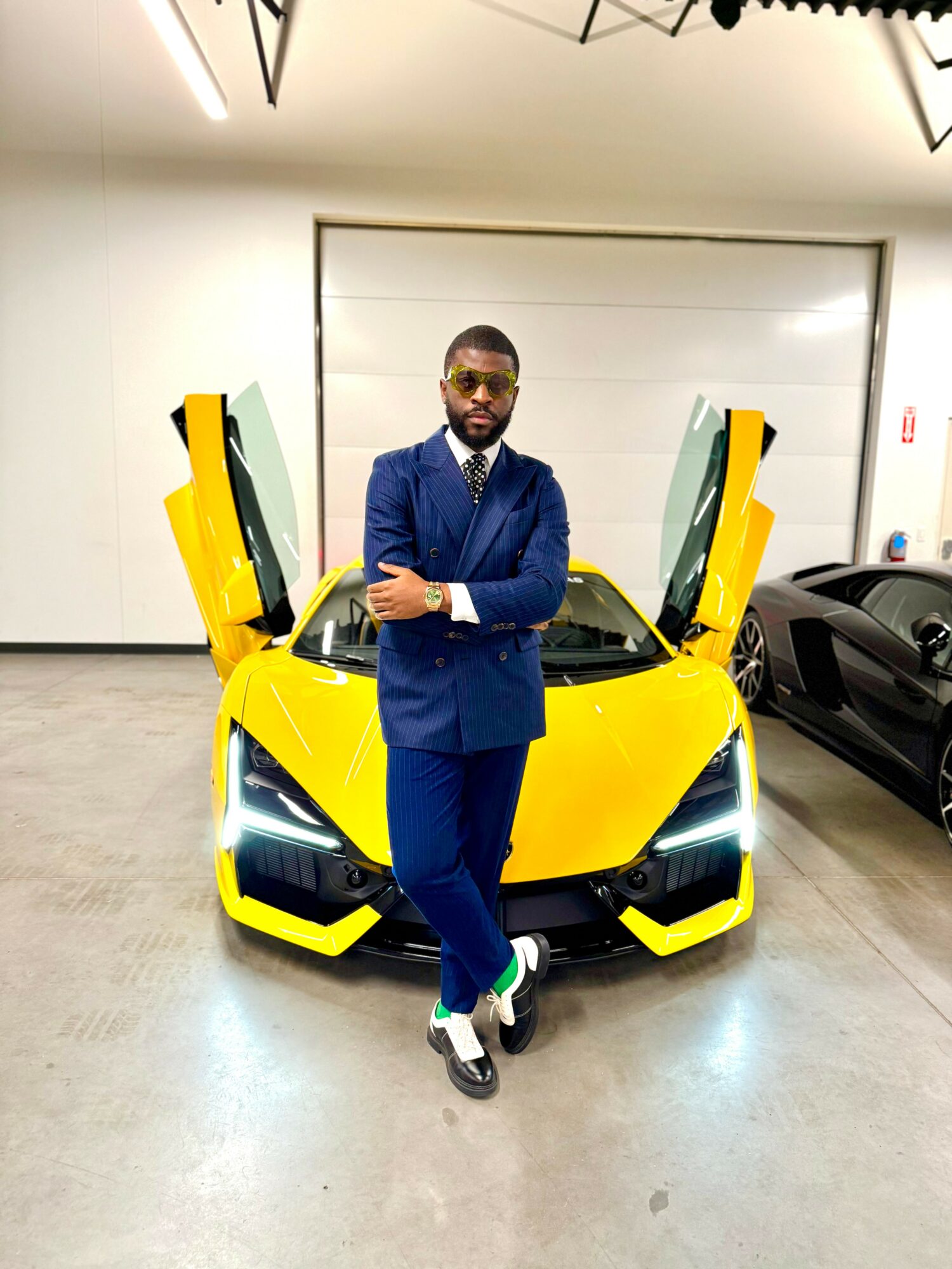 Man in a blue suit with crossed arms standing in front of a yellow sports car with open doors.