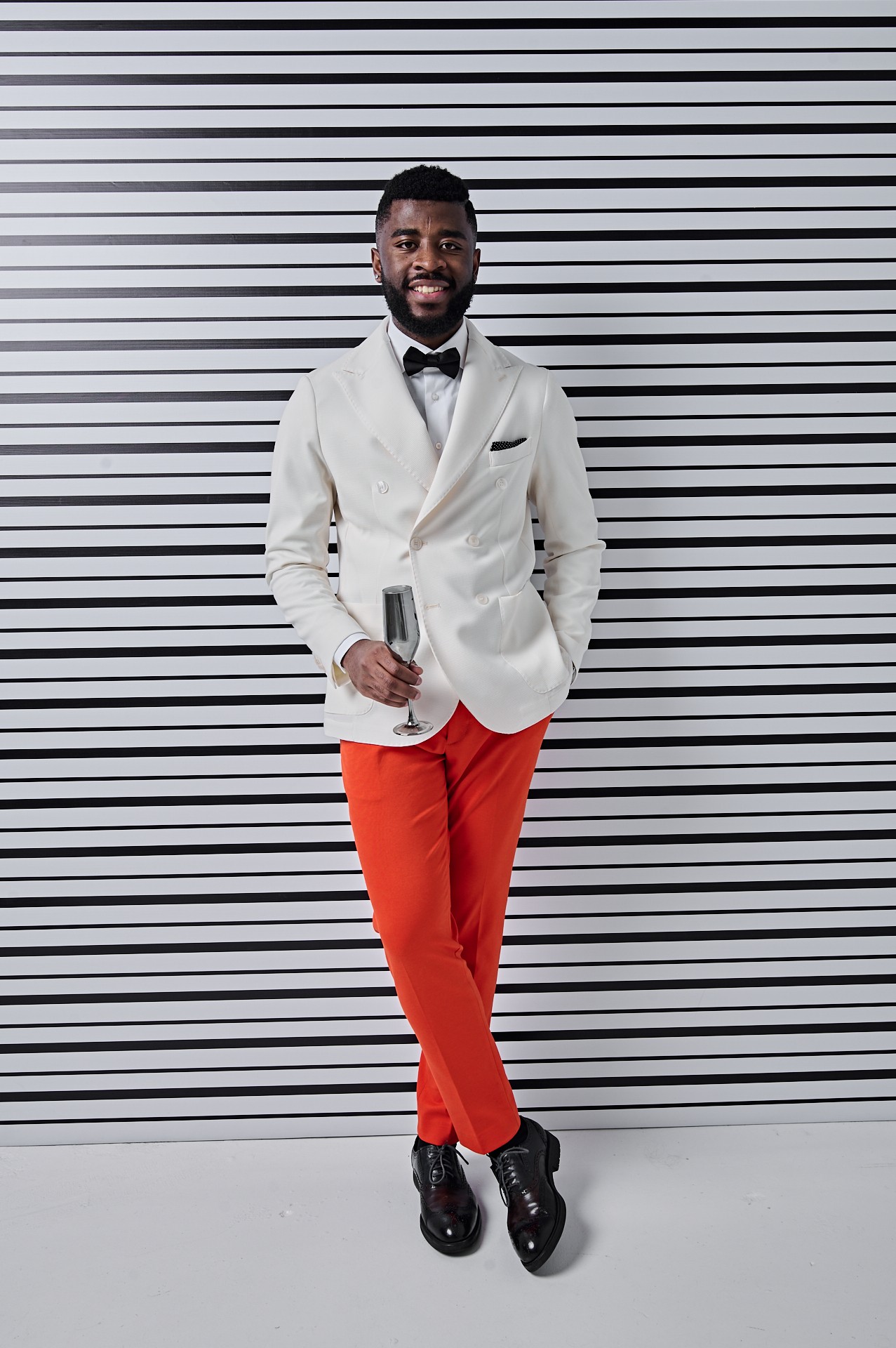 Man in tuxedo holding a glass, standing against a striped background.