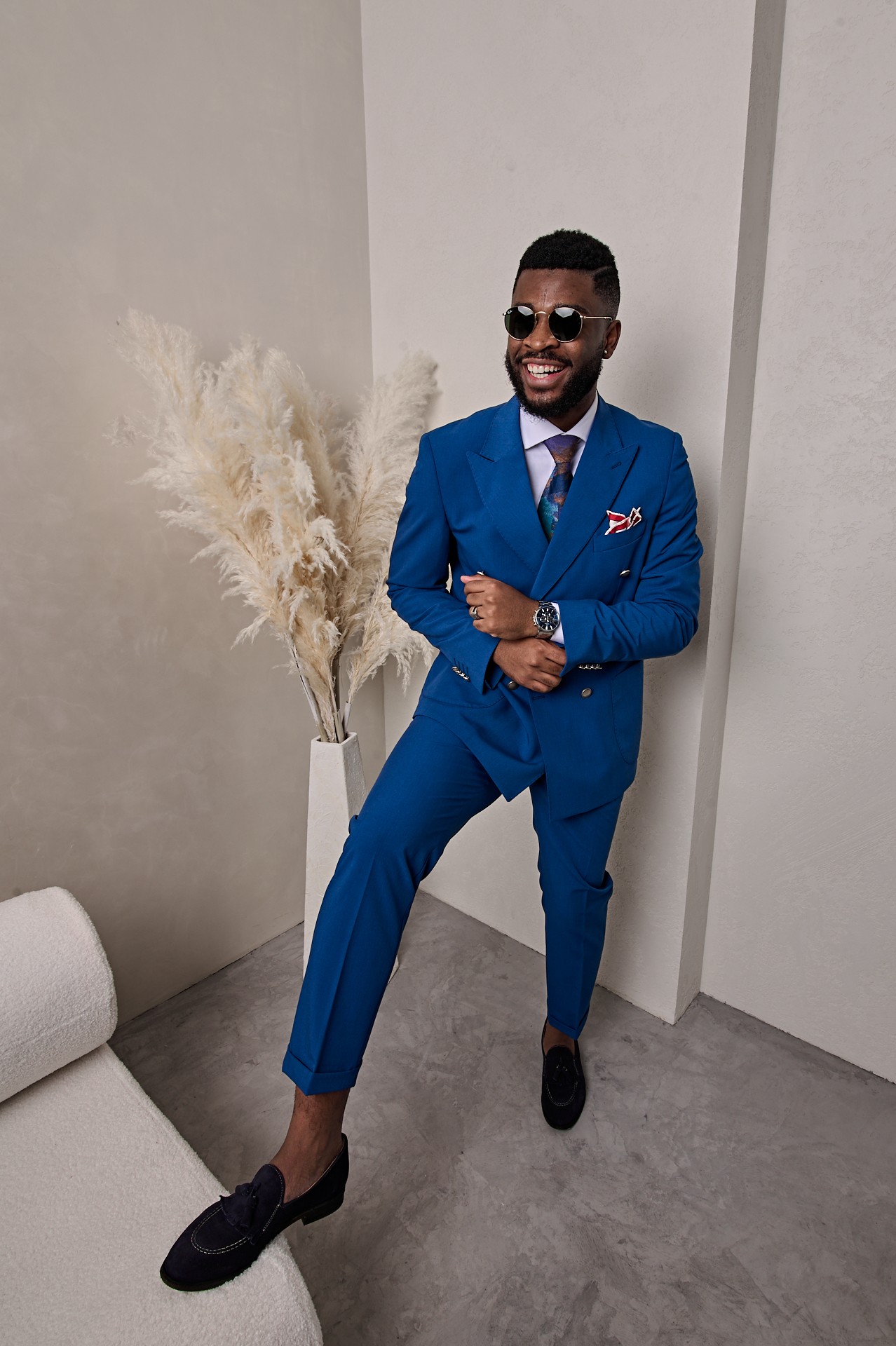 Man in a blue suit with sunglasses, smiling, standing indoors near a white wall and a vase with pampas grass.