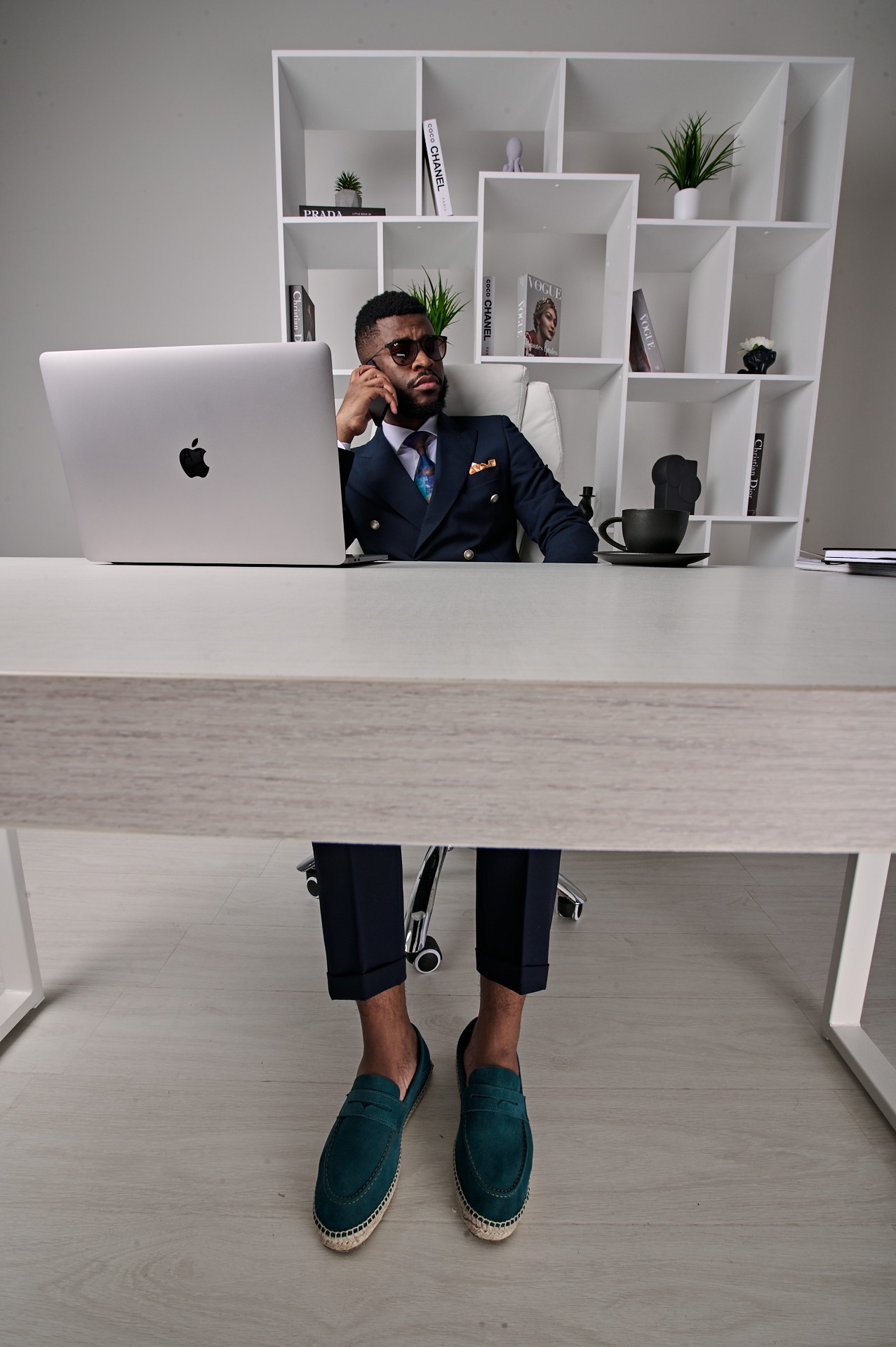 Man in suit sitting at desk with computer, bookshelf behind, in modern office space.