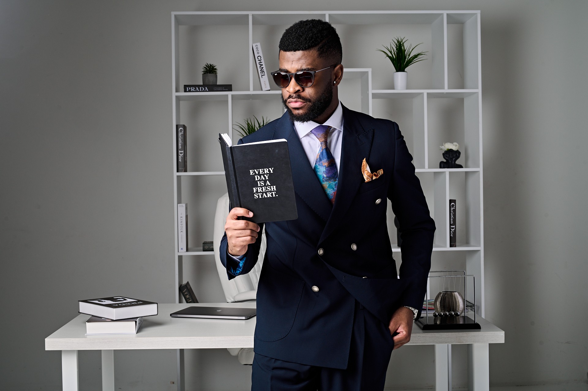 Man in a suit holding a black notebook, standing in front of a white bookshelf with decorative items.