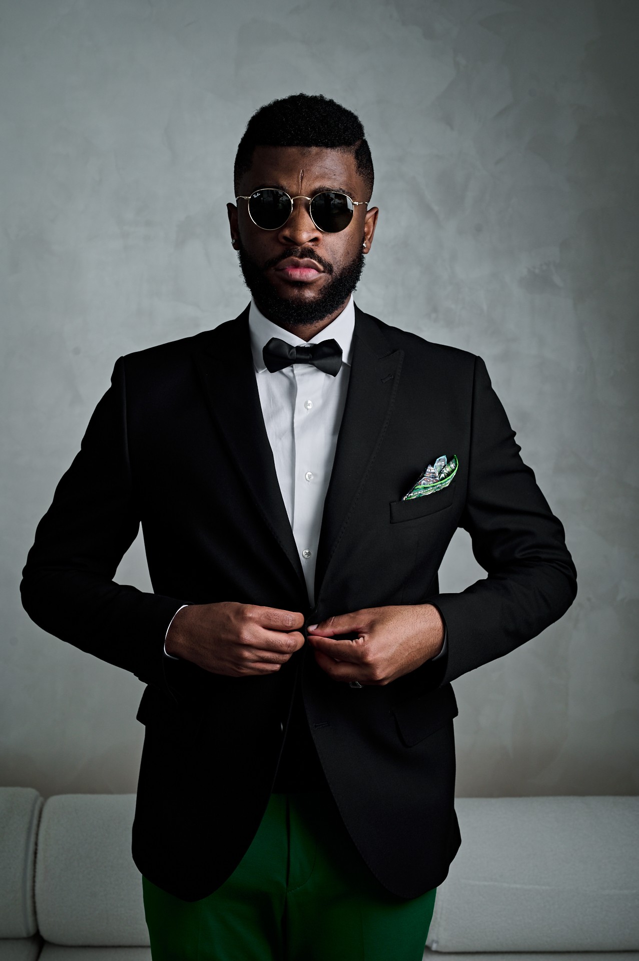 Man in black tuxedo with bow tie and sunglasses, standing against a gray background.