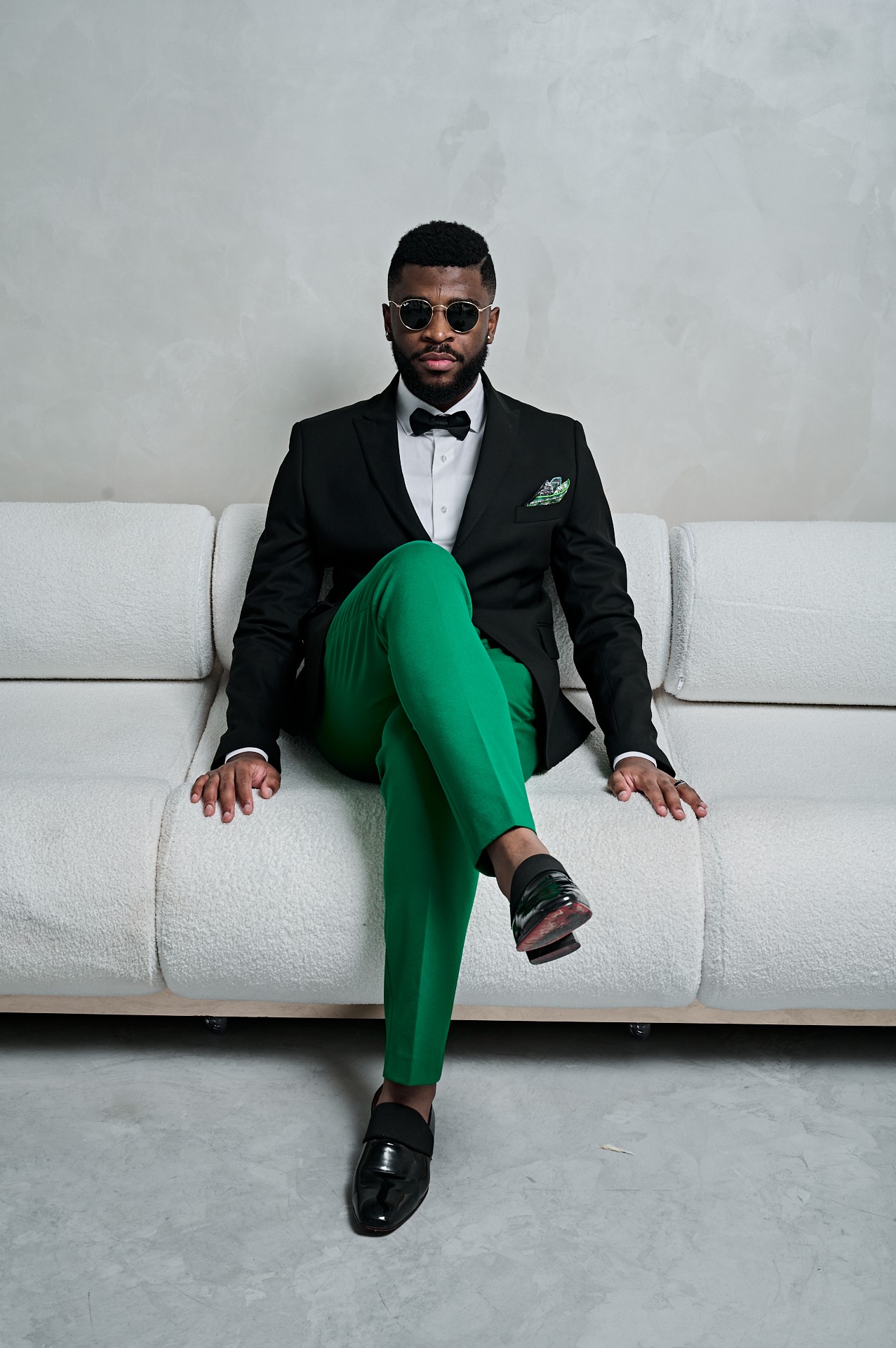 Man in tuxedo with green pants and sunglasses sitting on a white sofa against a plain wall.