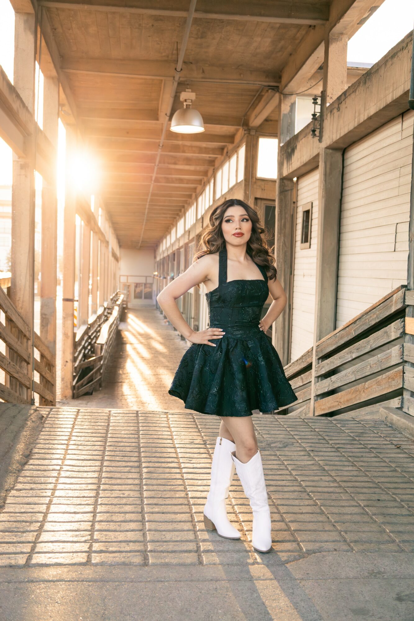 Woman in black dress and white boots standing on wooden walkway with sunlight shining behind her.