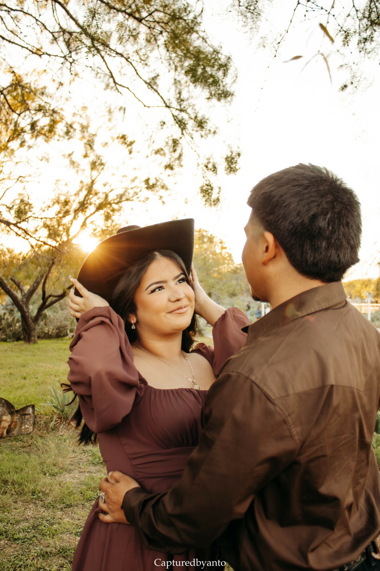 A woman and man outdoors during sunset, facing each other, with trees and sky in background.