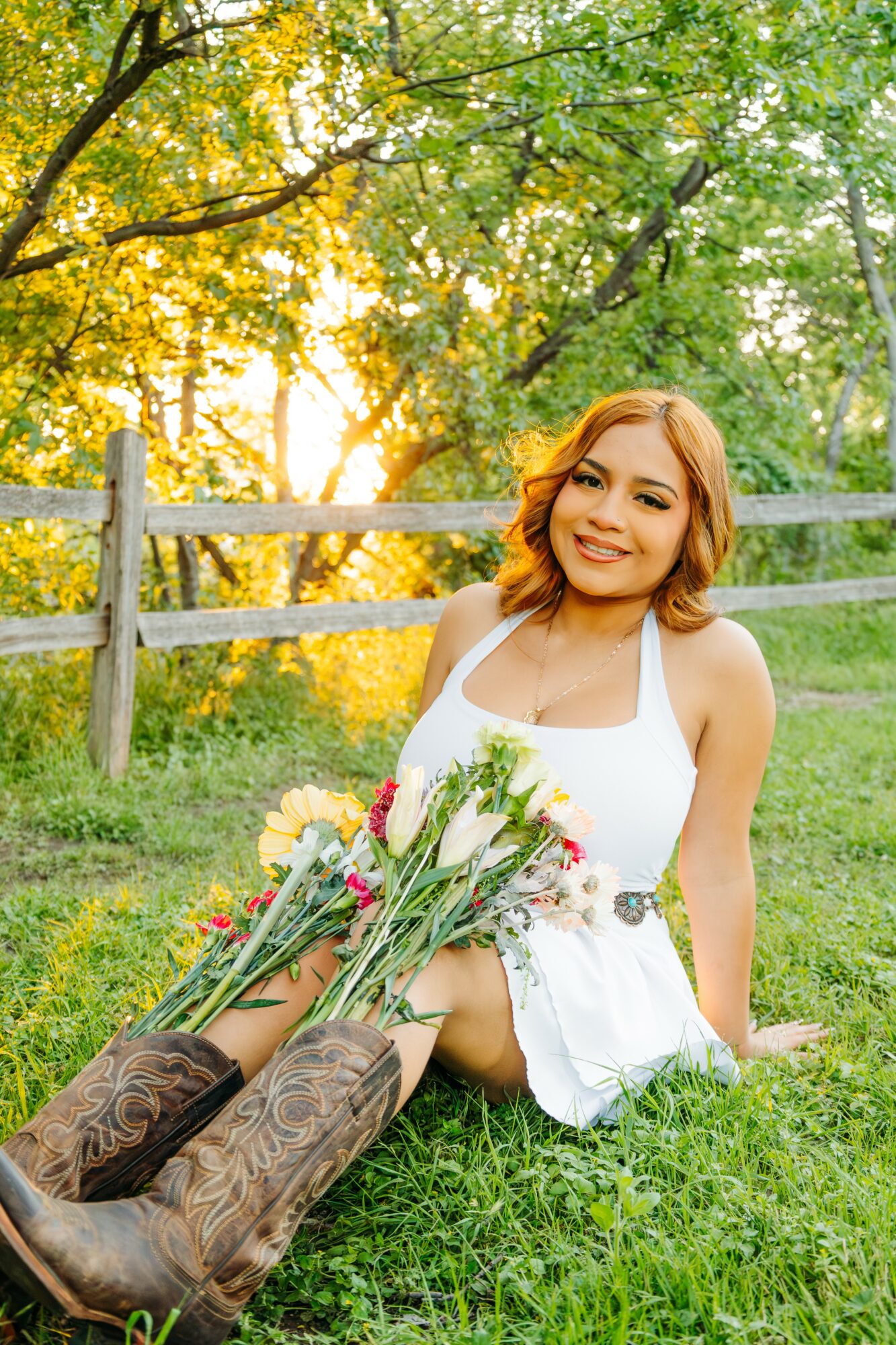 Young woman with shoulder-length hair smiling, sitting on grass with a bouquet of flowers, outdoors with trees and a fence.