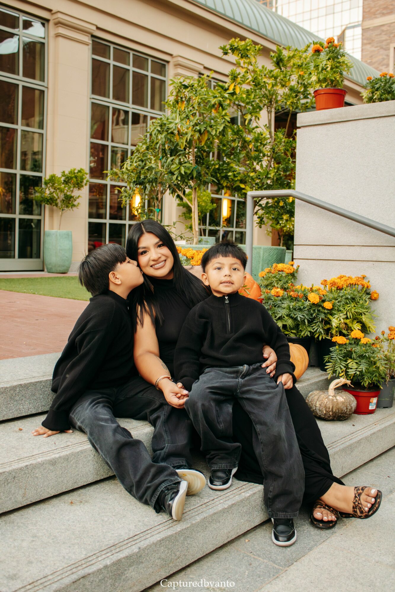 Woman with two children sitting on outdoor steps surrounded by potted plants and flowers.