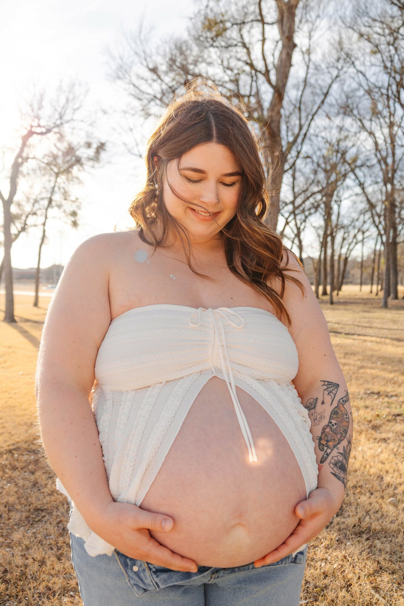 Pregnant woman outdoors, holding her belly, smiling, with trees and a field in the background.