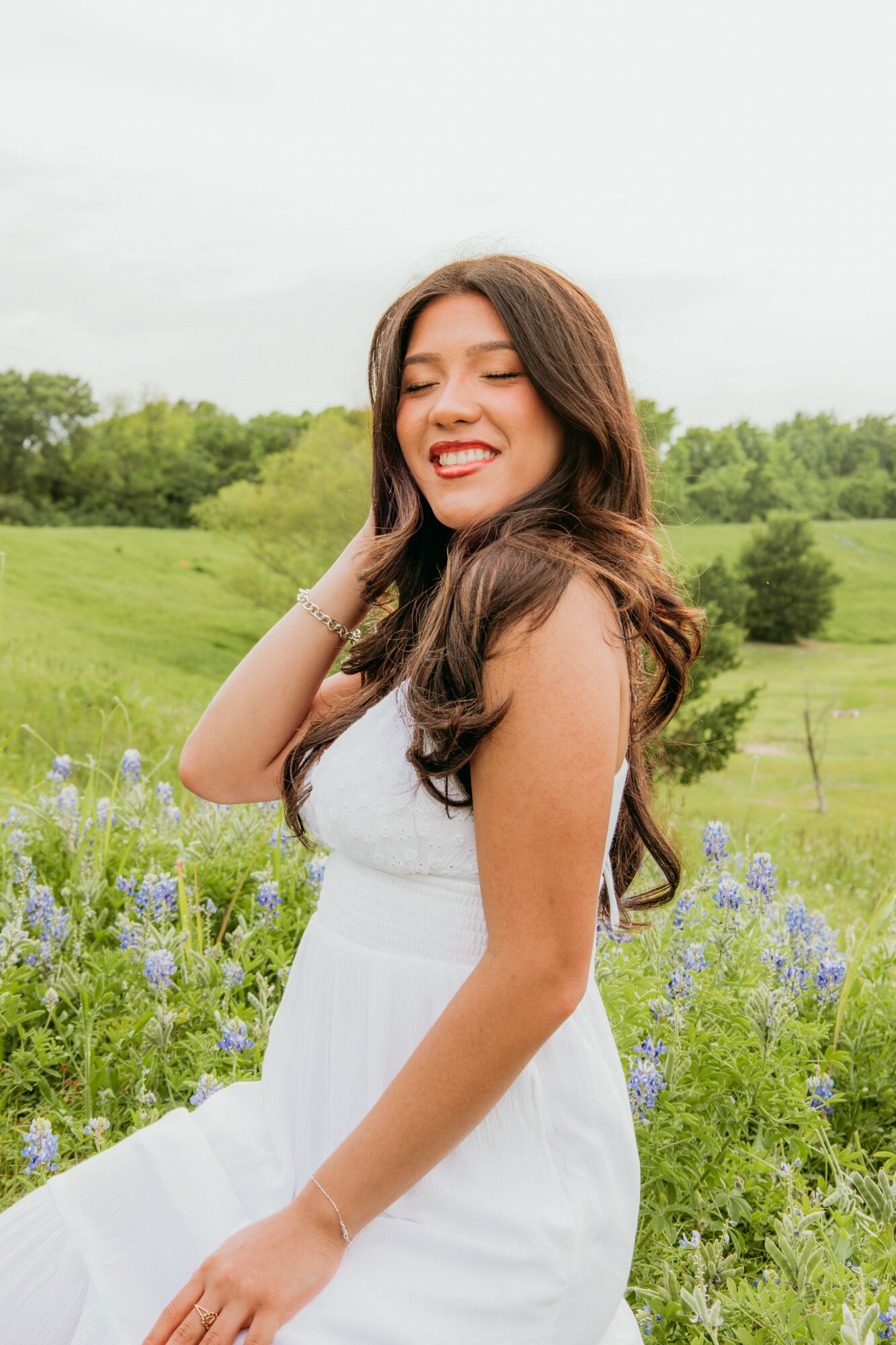 Young woman with long hair smiling outdoors in a field of purple flowers and green trees.