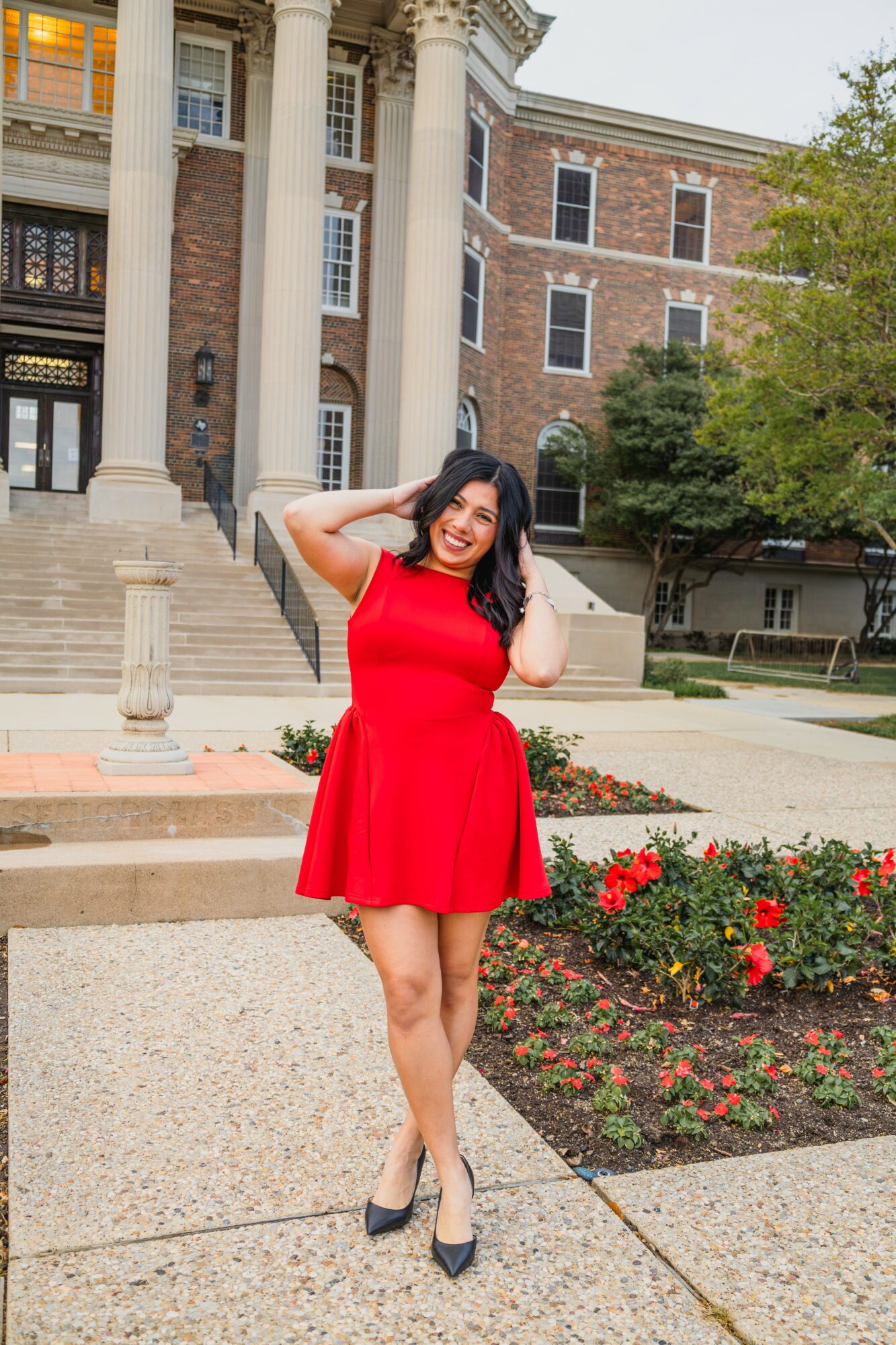 Young woman in red dress standing outdoors near flower bed, smiling, with historic building in background.