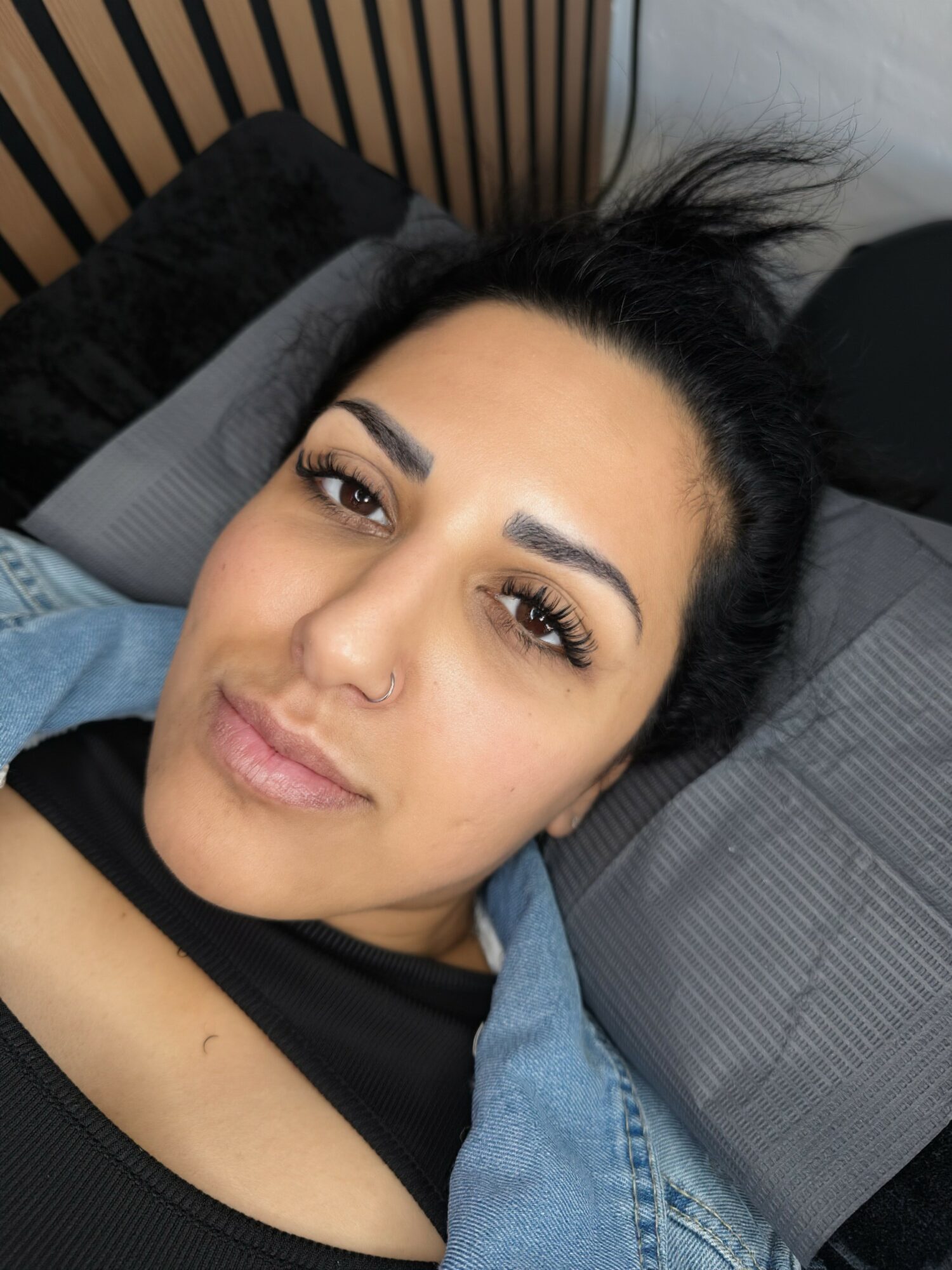 Woman with dark hair lying on bed, looking at camera, with pillows and headboard visible.