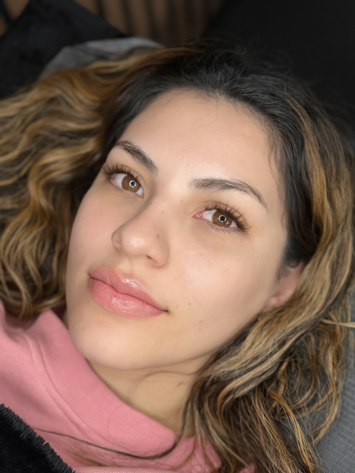Close-up of a woman with wavy hair, lying down, wearing a pink top, looking at the camera.