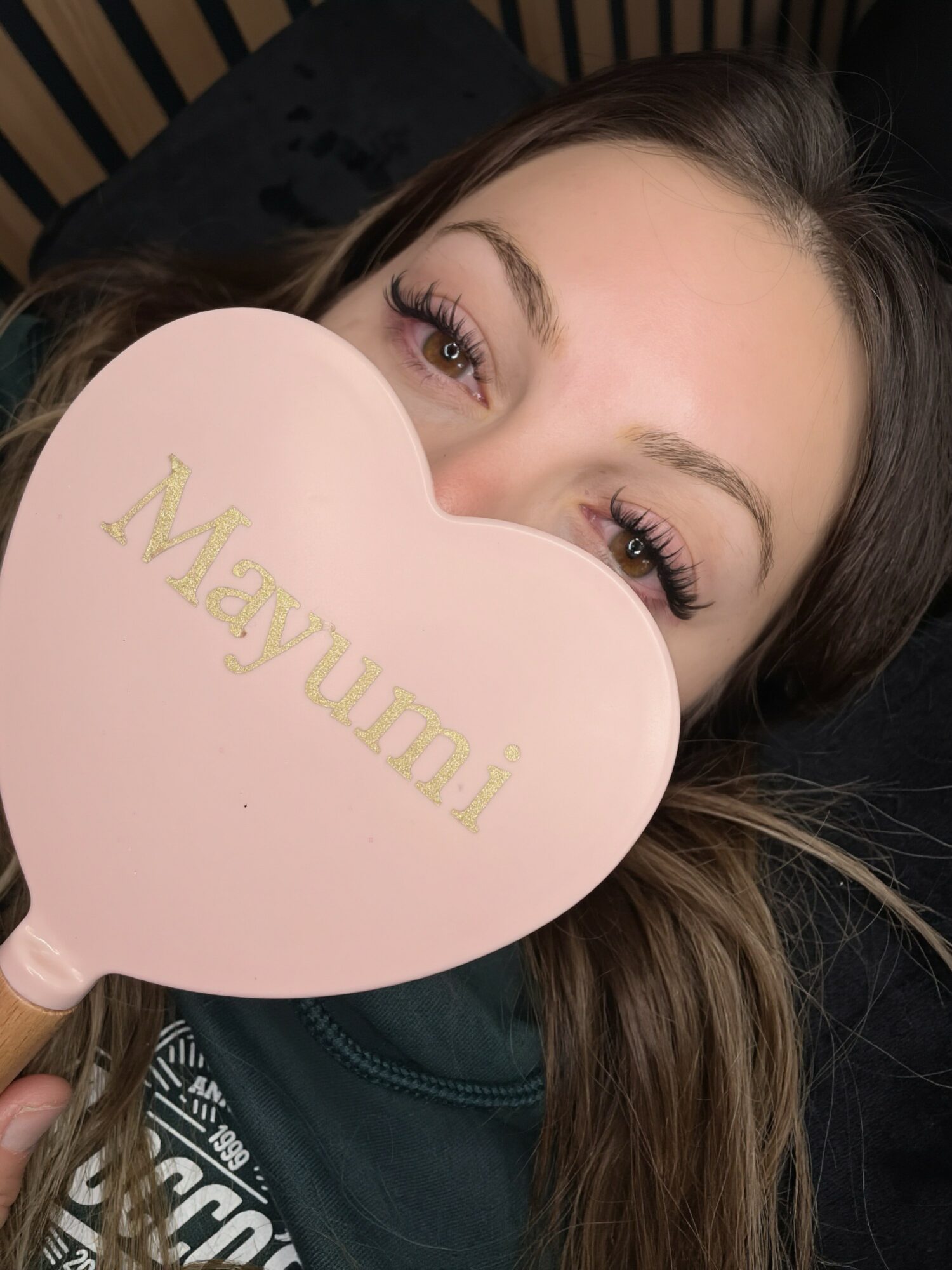 Person holding a pink heart-shaped sign with gold text partially covering face, eyes visible, dark background.