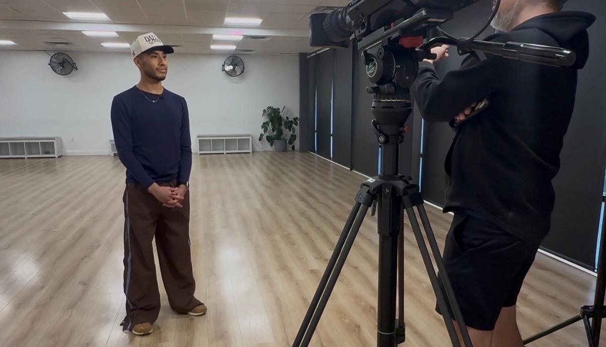 Man standing in a large, empty room being filmed by a person with a camera on a tripod.