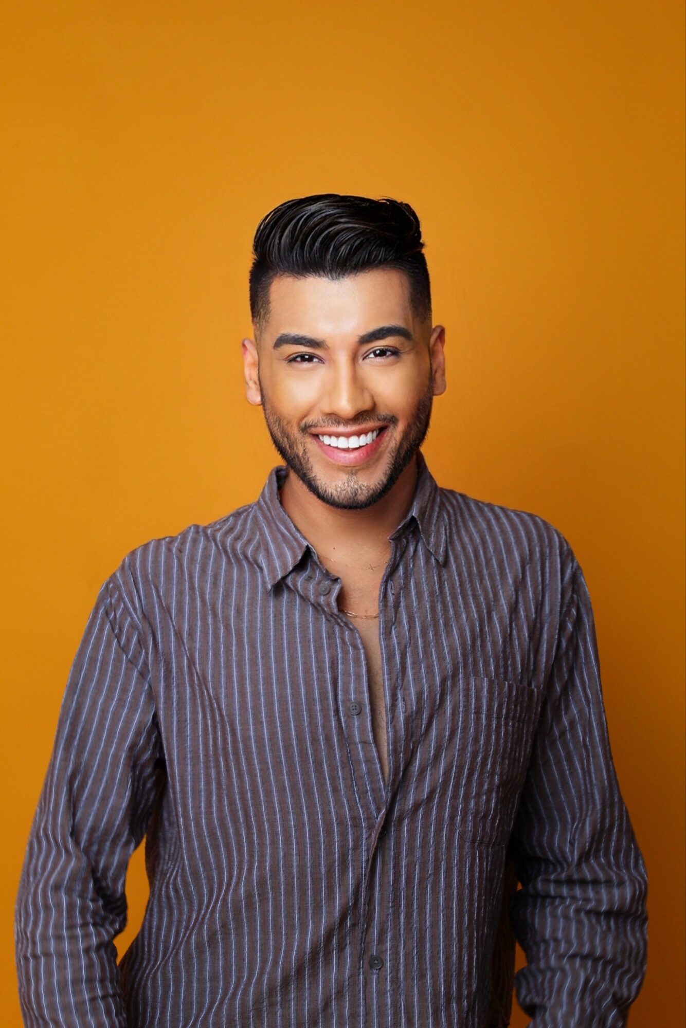 Smiling man with dark hair and a striped shirt against a yellow background.