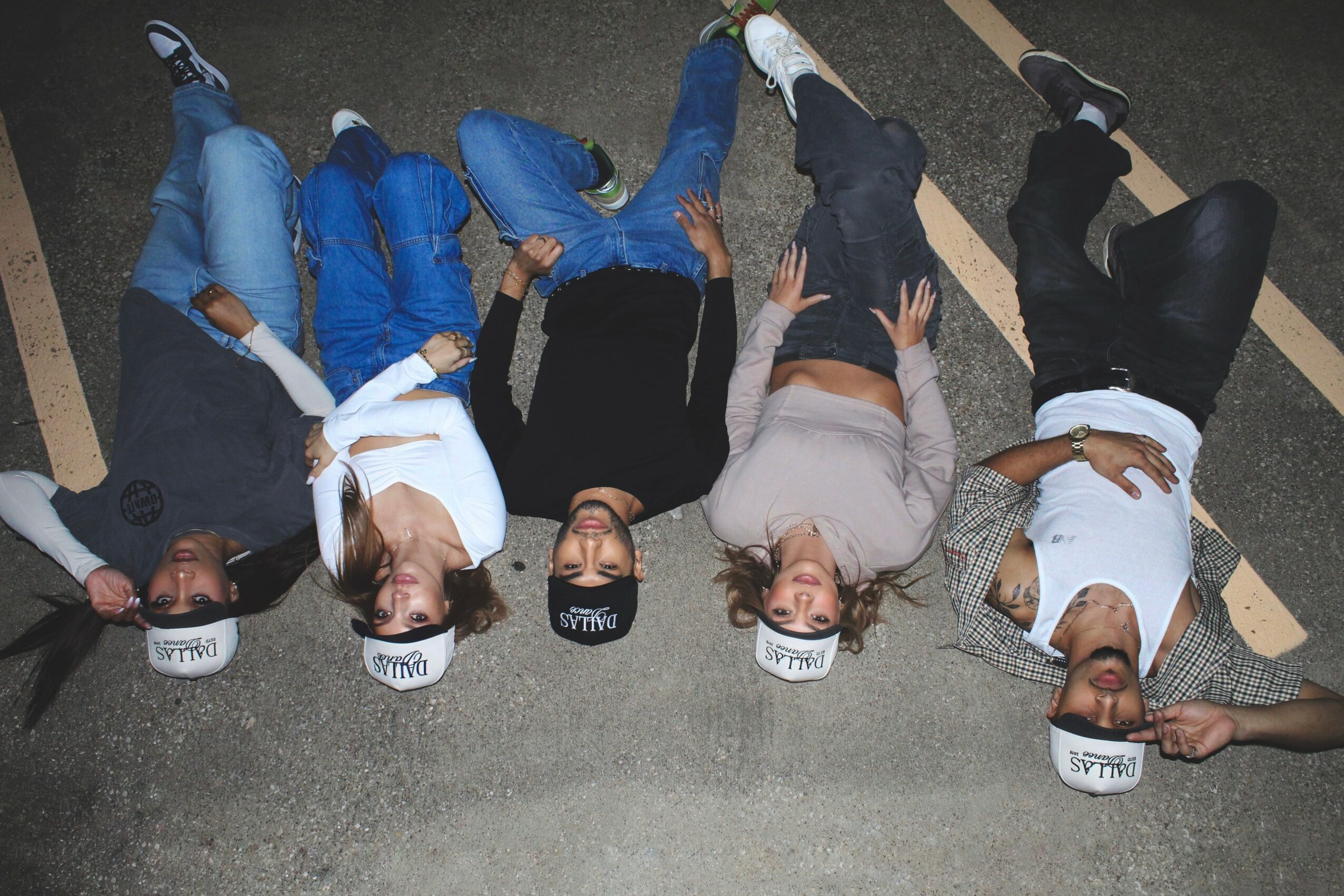 Group of five people lying on the ground in a parking lot, wearing white hats and casual clothing.
