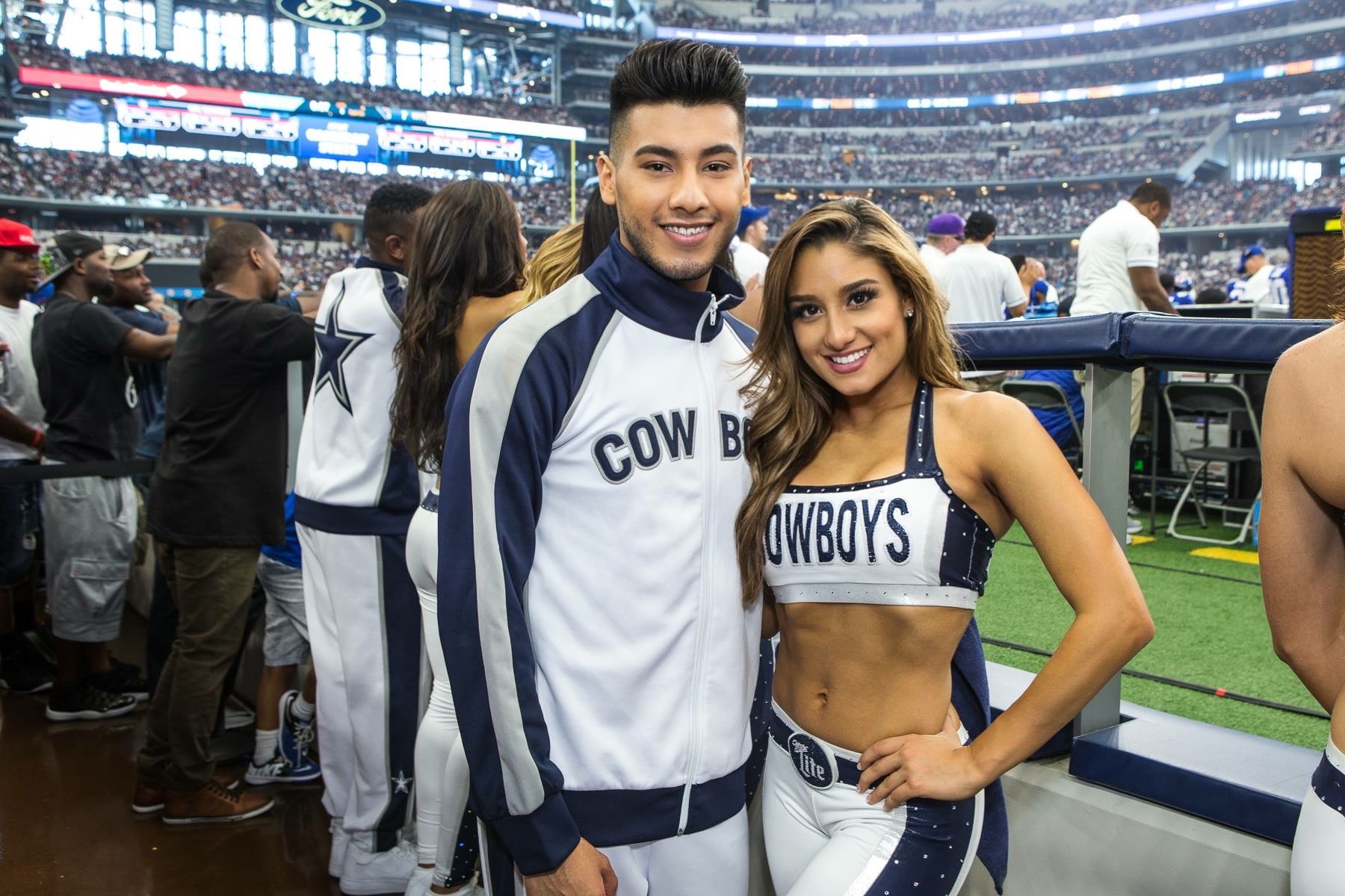 Two smiling people in Dallas Cowboys apparel at a stadium, with a crowd in the background.