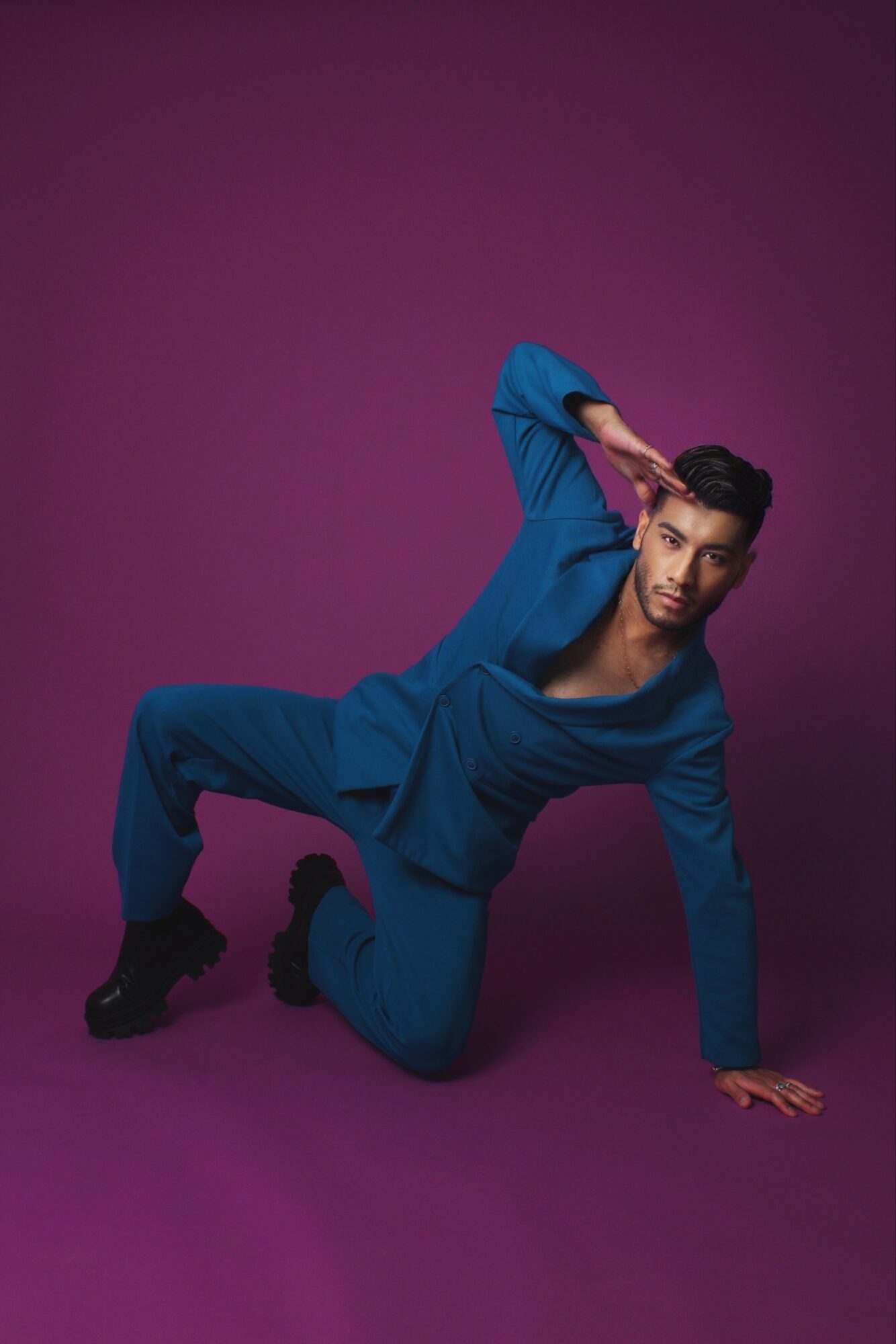 Person in blue outfit crouching on purple background, one hand on the ground, other hand near head.