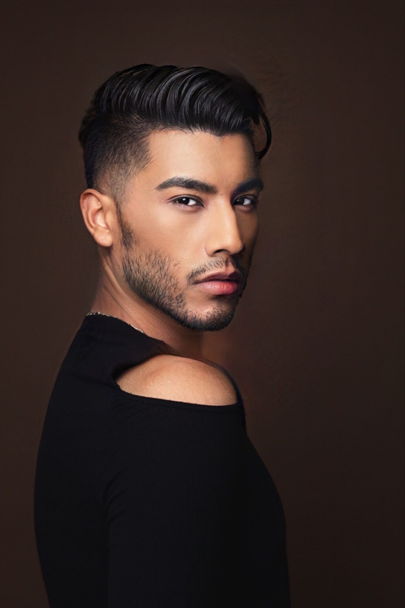 Young man with styled hair and beard, wearing a black top with a shoulder cutout, looking sideways.