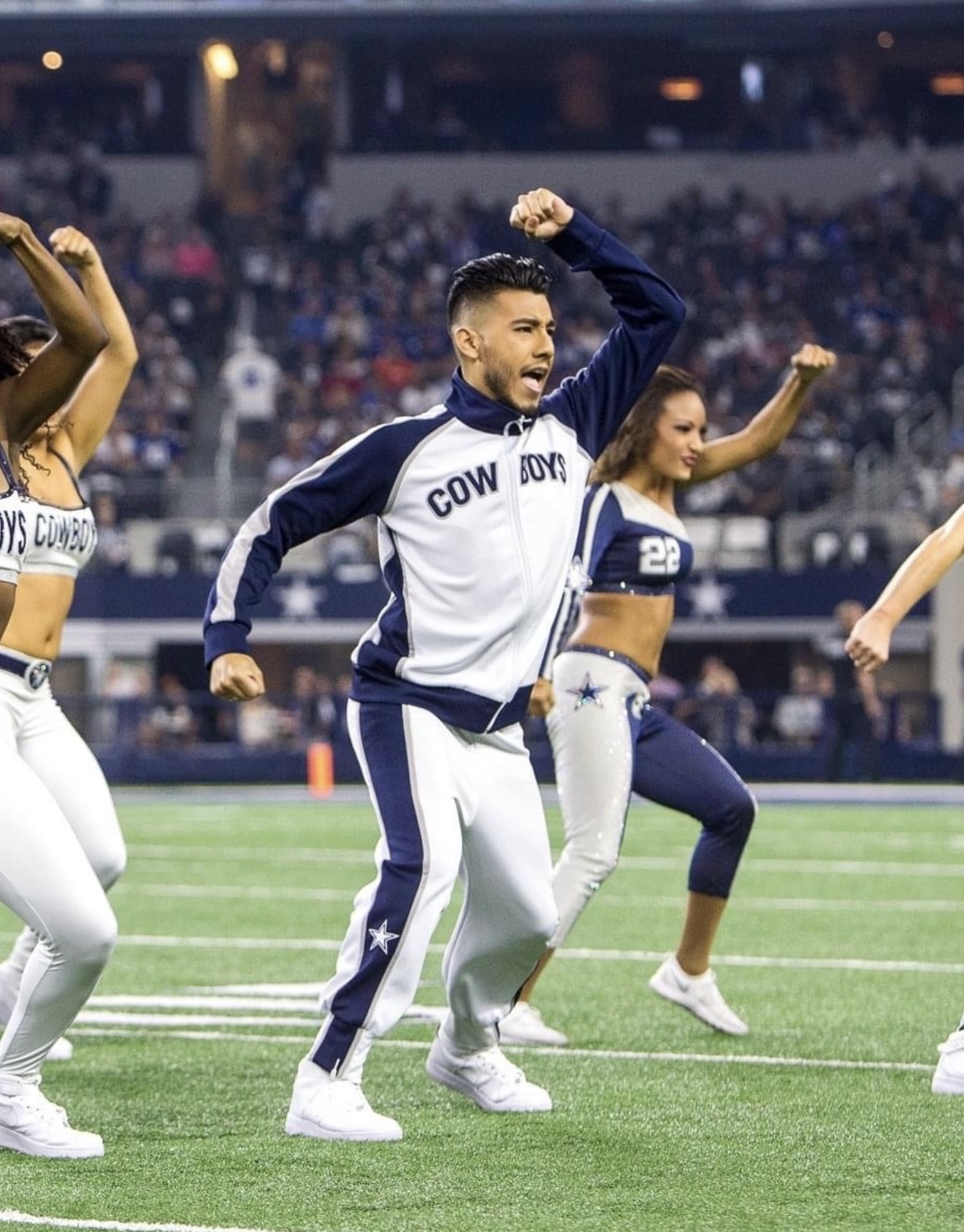 Football player celebrating with raised fist on field, other players and stadium in background.