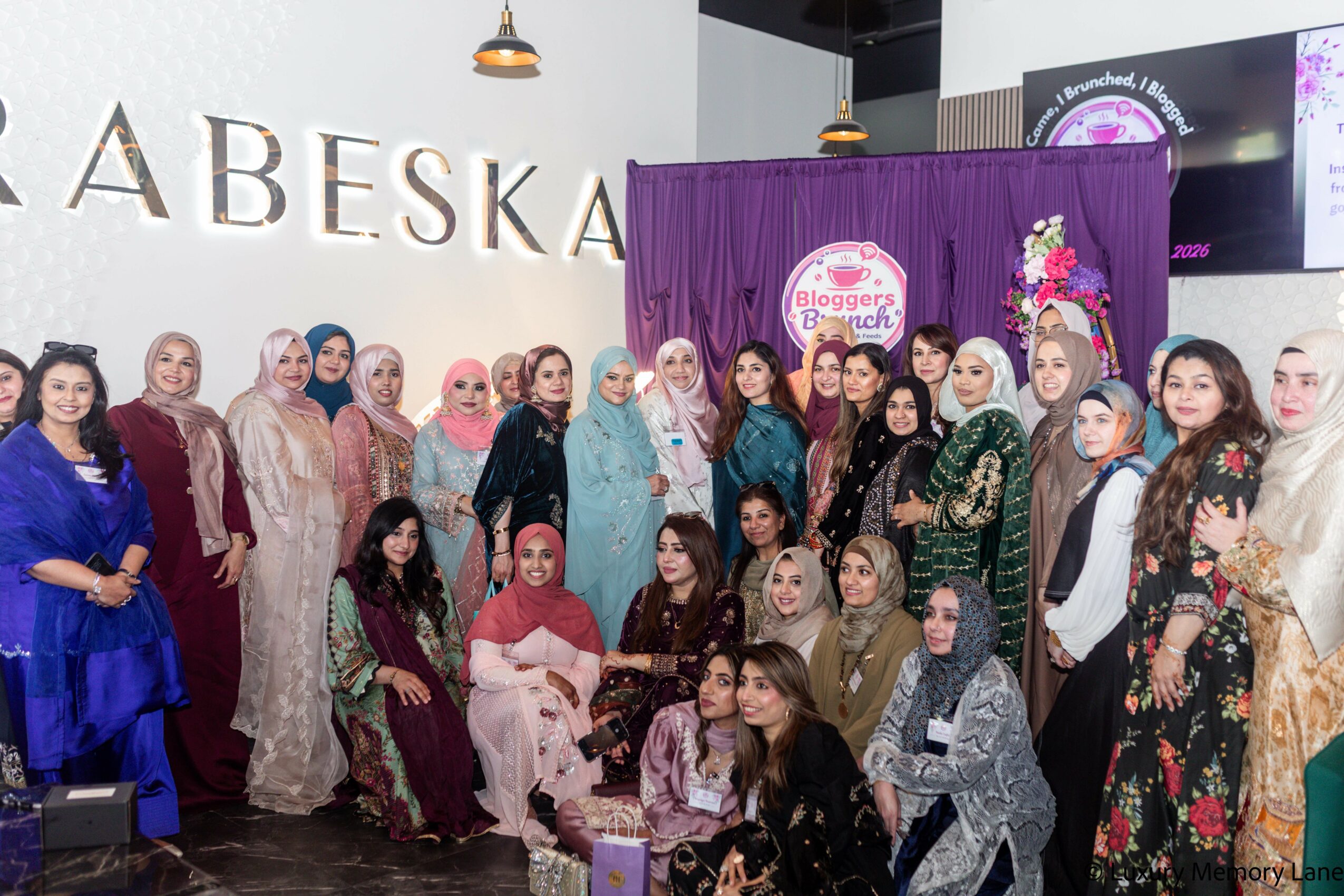 Group of women and girls standing in front of a purple backdrop with a logo, indoors, some wearing traditional clothing.