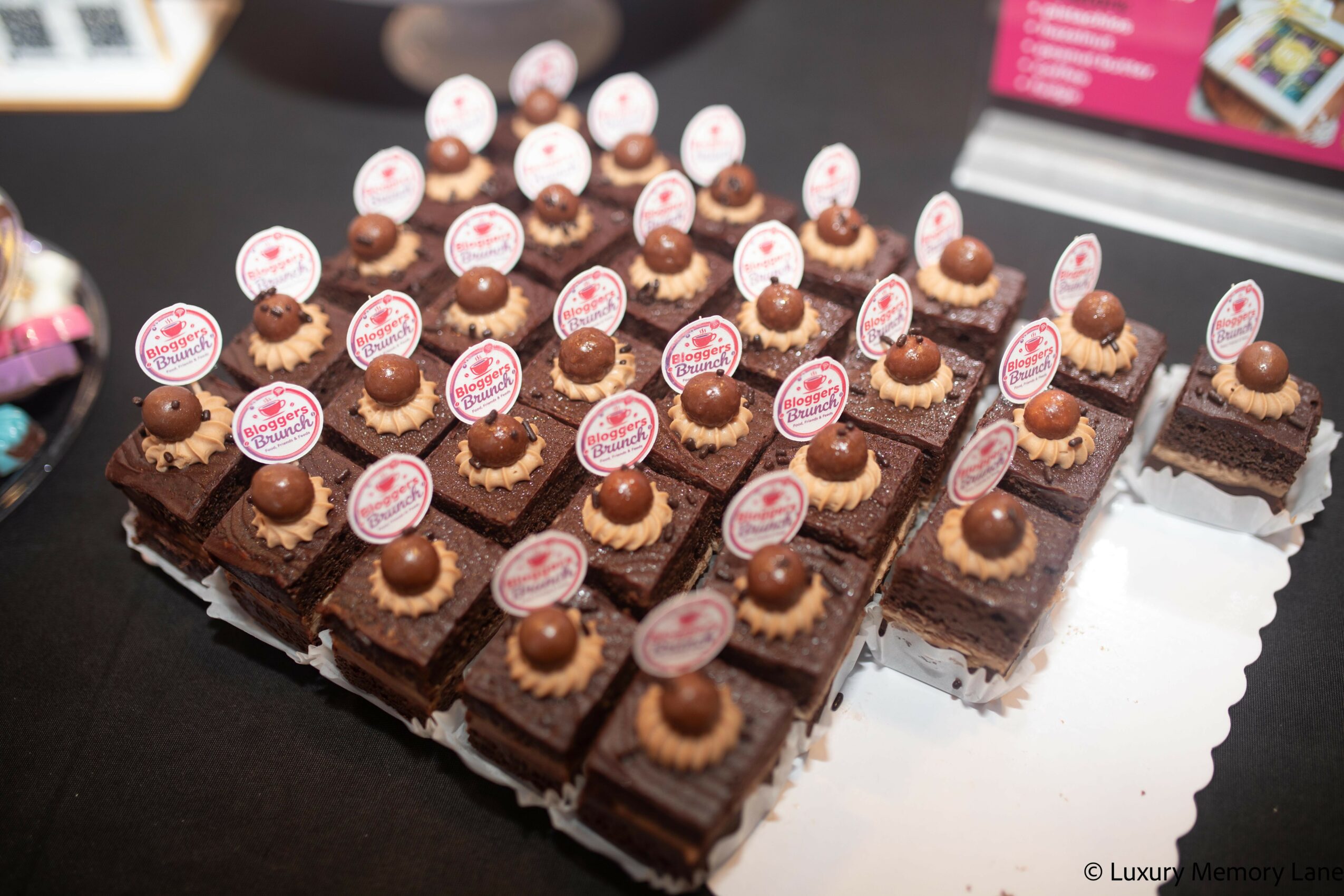 Assorted chocolate desserts with decorative toppings on a reflective surface, some with small round labels, arranged in rows.