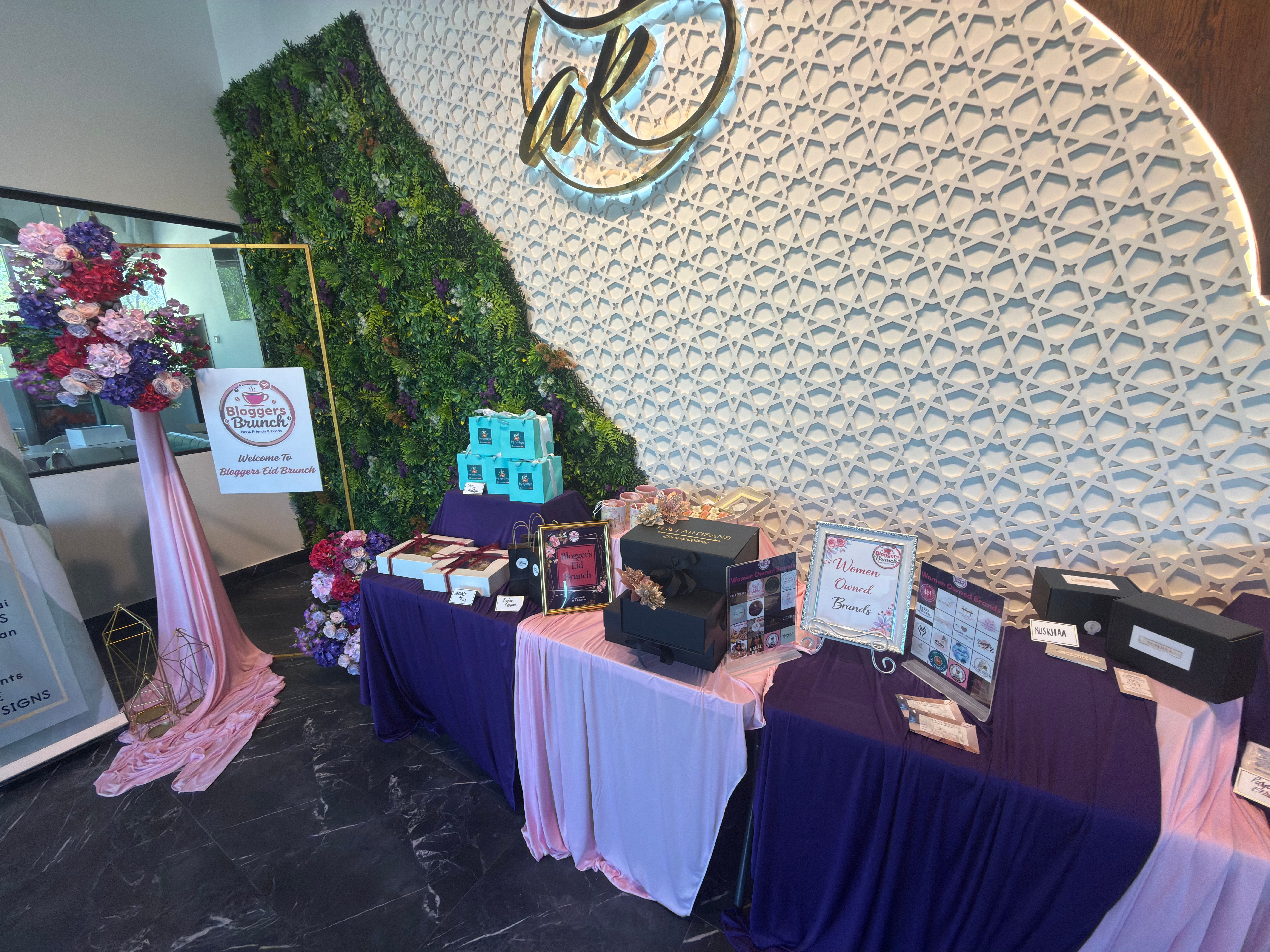 Table with gift boxes and framed signs, decorated with purple and white cloths, against a textured wall and greenery backdrop.