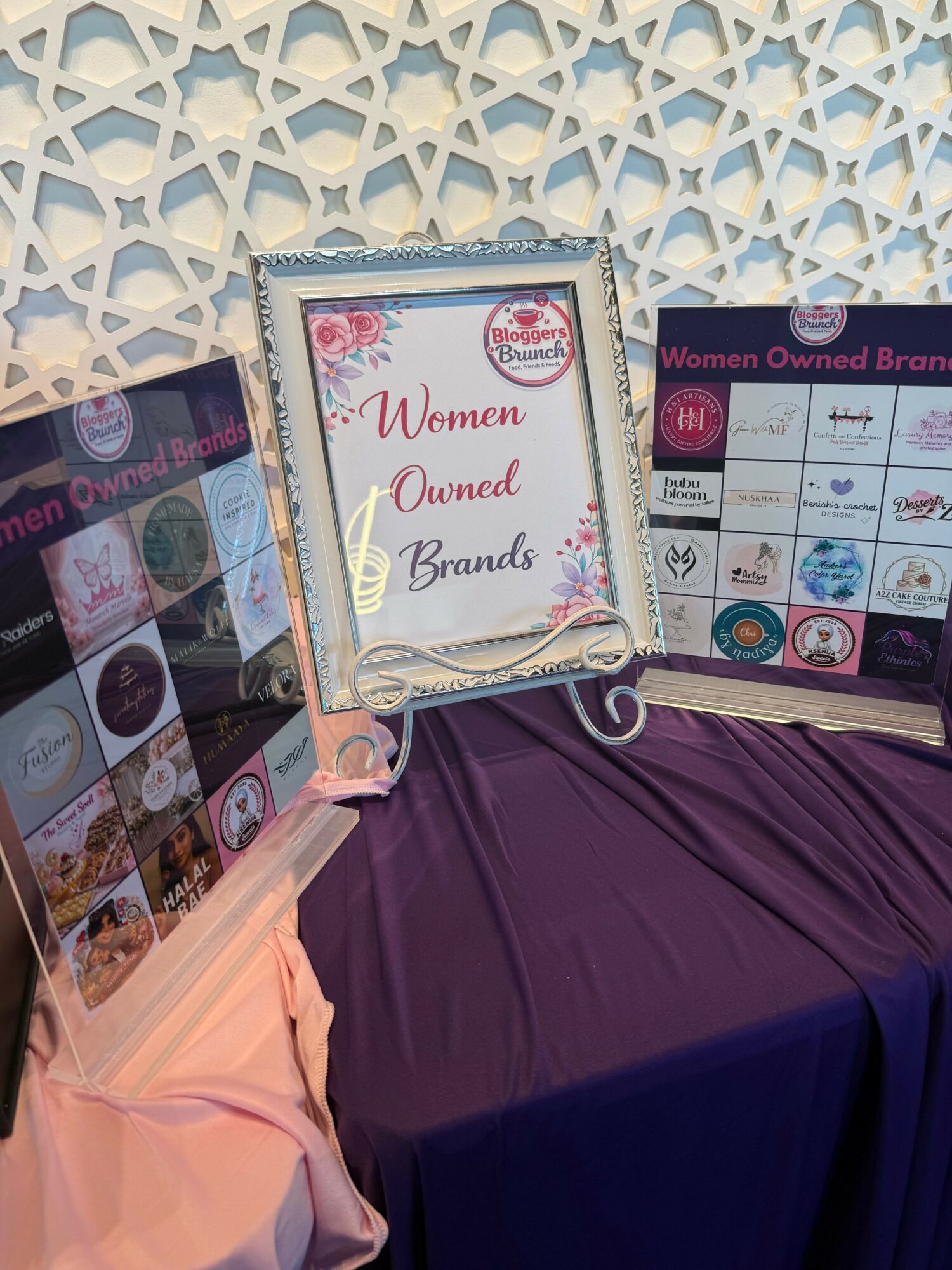 Display table with framed sign reading 'Women Owned Brands' and promotional materials, draped with purple cloth, against decorative background.