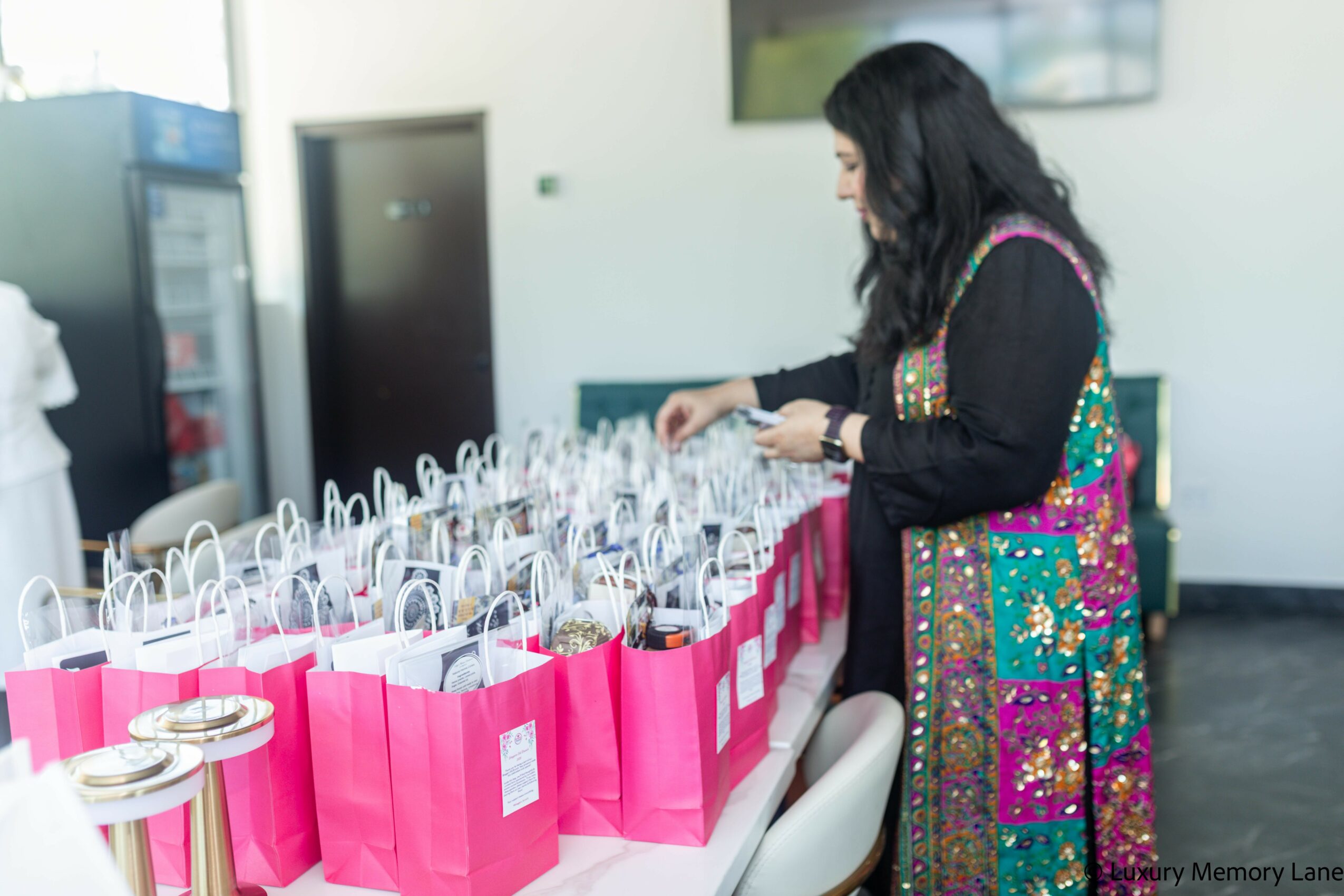 Woman with long dark hair wearing a colorful dress, standing at a table with pink gift bags.