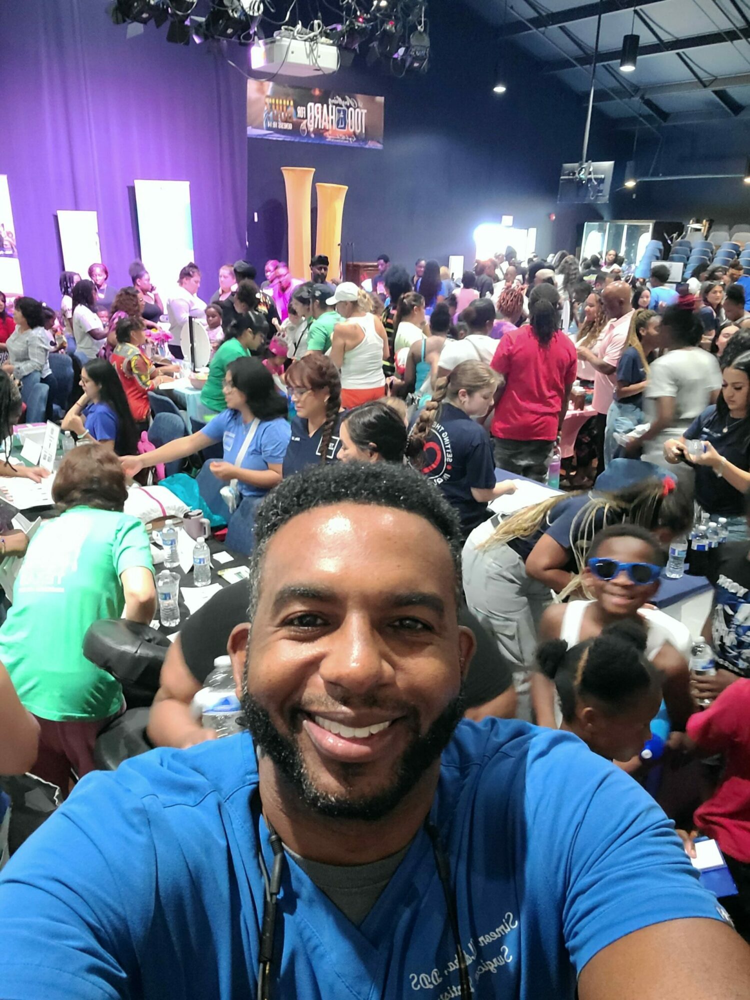 Man smiling in a crowded indoor event with many people seated and standing, colorful lighting, and a stage in the background.